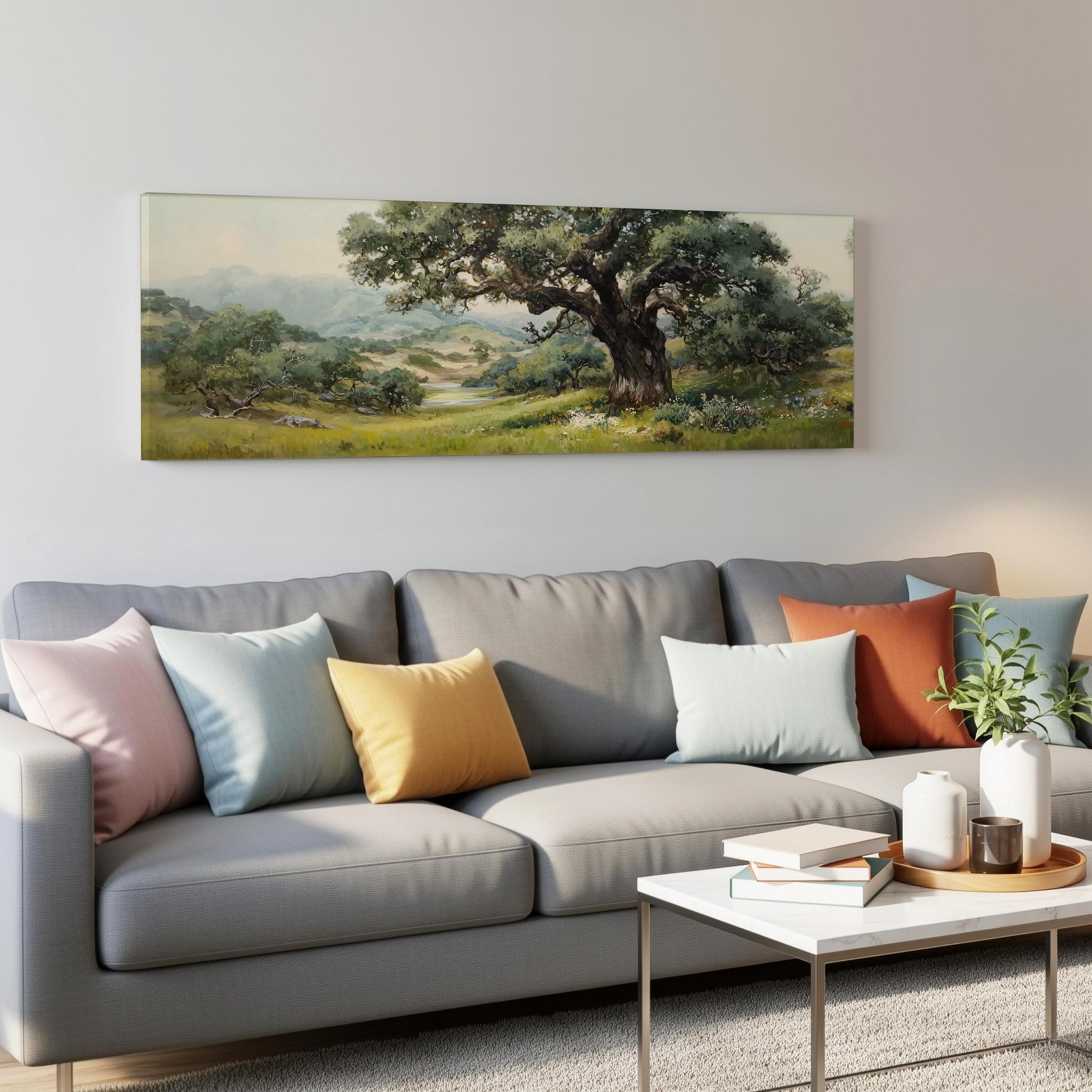 a living room with a gray sofa adorned with colorful throw pillows, a coffee table with books and a plant, and a large landscape painting hanging on the wall above the sofa.