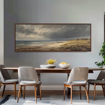 a dining room with a large painting of a beach scene hanging on the wall above a wooden dining table with four chairs. The painting depicts a cloudy sky over the ocean, with a sandy beach and grassy hills in the background.