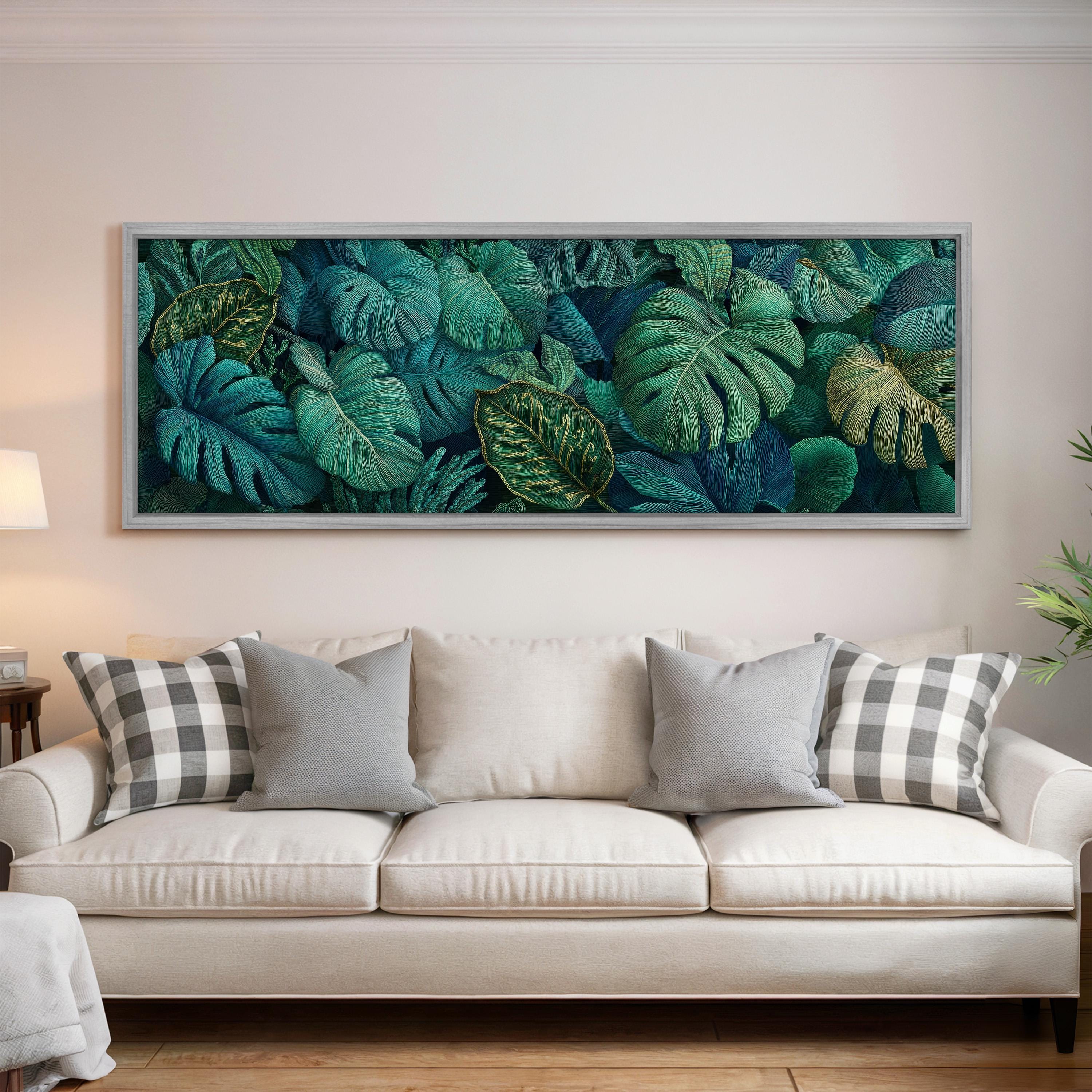 a large framed painting of tropical leaves hanging on a wall above a white sofa with gray and white patterned pillows.