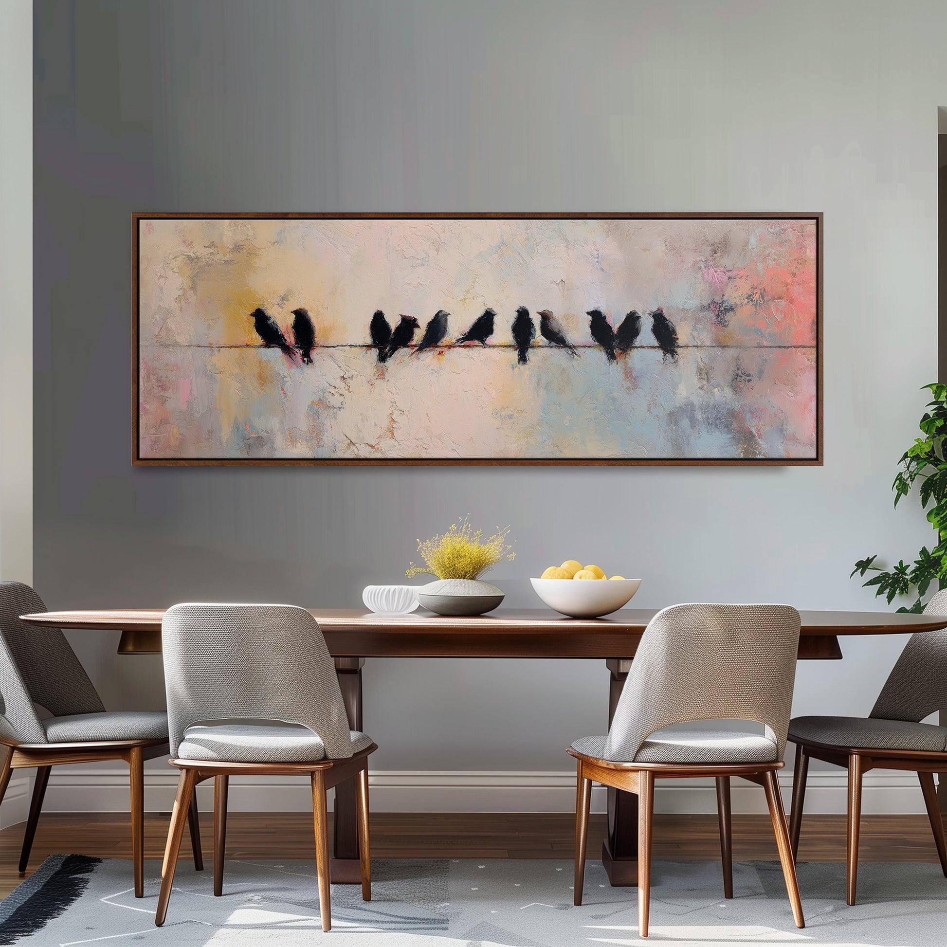 a dining room with a large painting of birds perched on a wire above a wooden table. The table is set with a bowl of fruit and a vase of flowers. The room has a neutral color scheme with gray walls and wooden furniture.