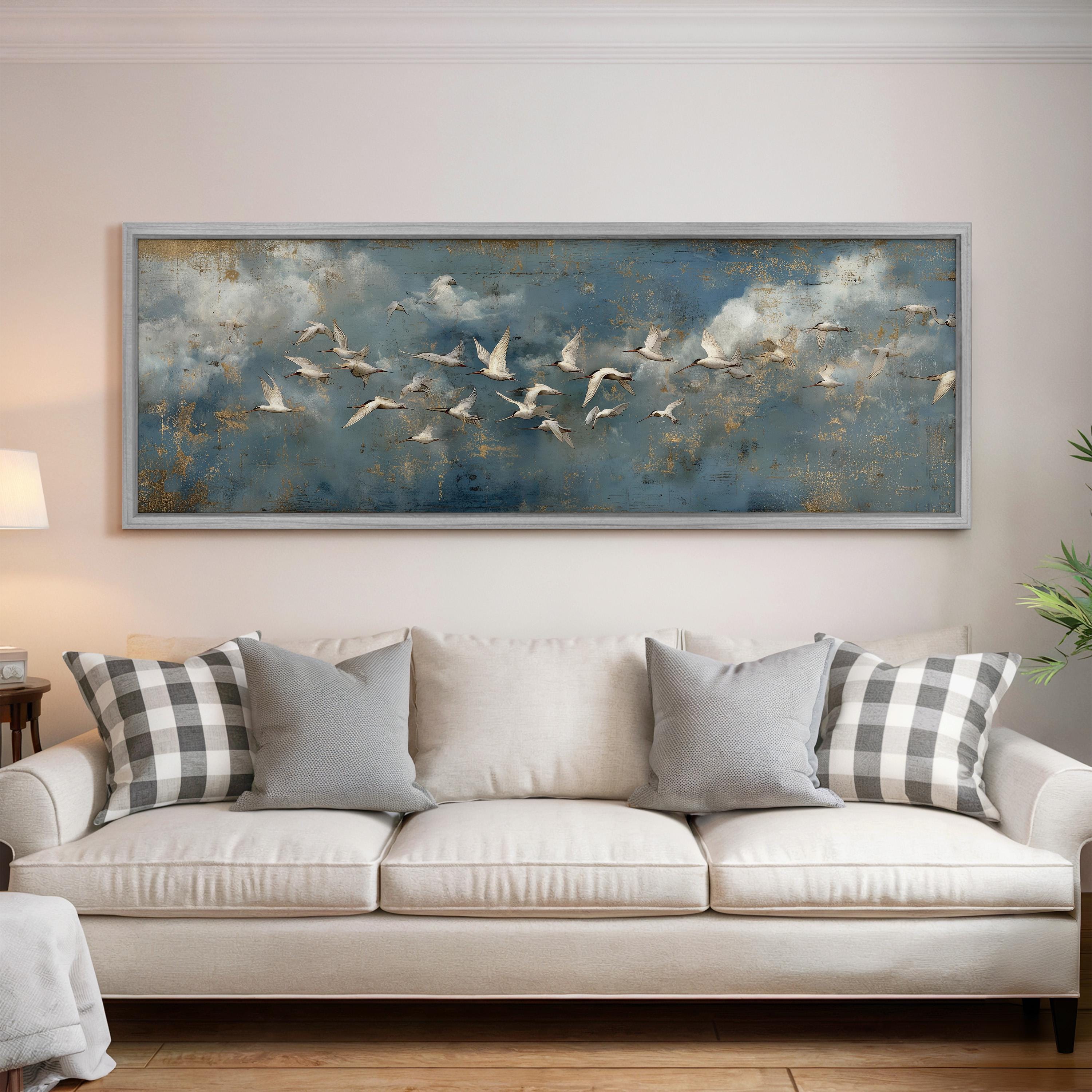 a large painting of a flock of birds flying over a cloudy sky, hanging on a wall above a white sofa with gray and white patterned pillows.