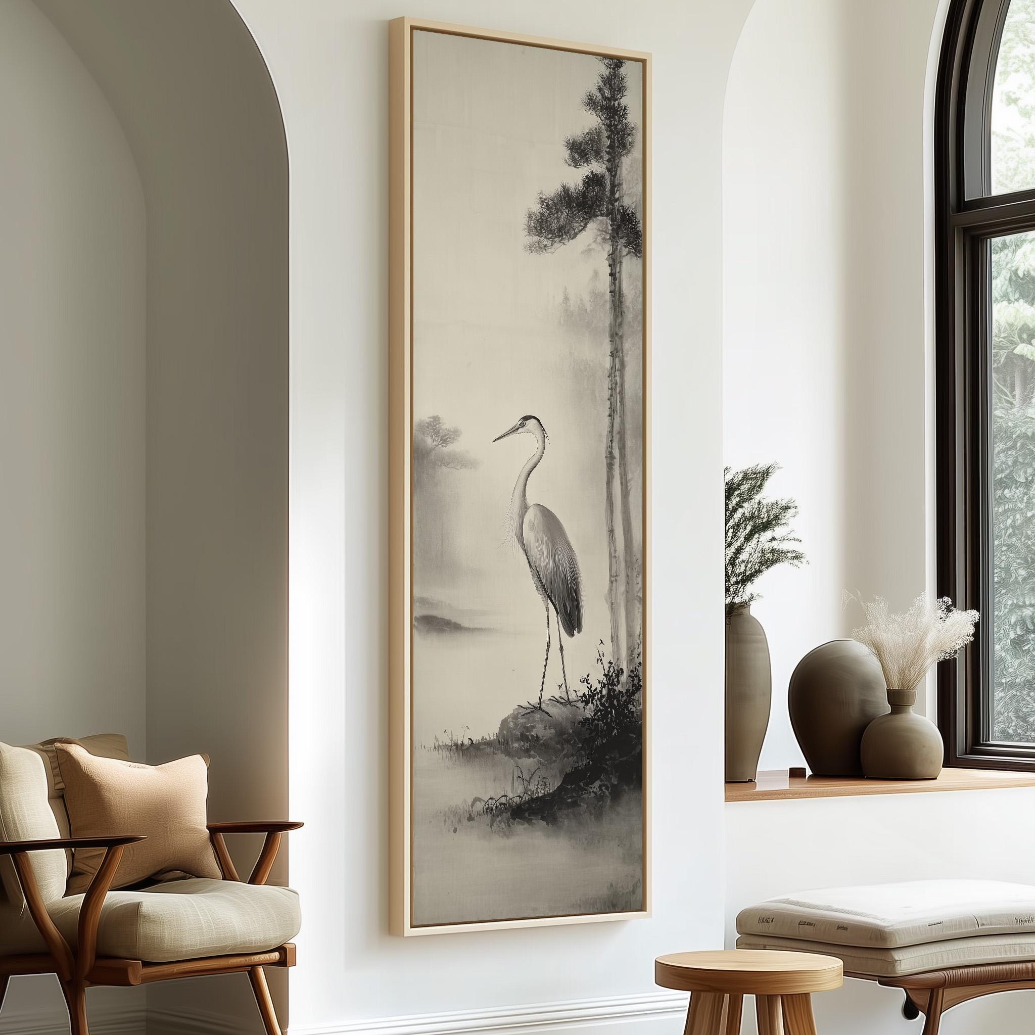 a large black and white painting of a crane standing on a rock, hanging on a wall in a room with a window and a wooden chair.