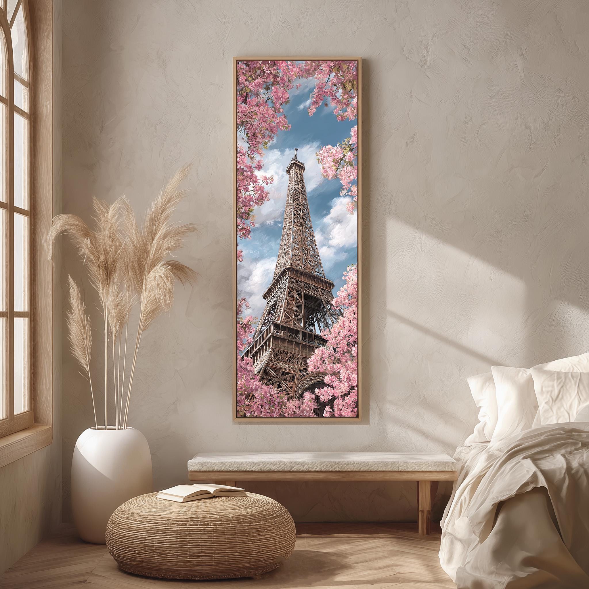 a large framed picture of the Eiffel Tower, surrounded by pink flowers, hanging on a wall in a room with a bed, a potted plant, and a woven ottoman.