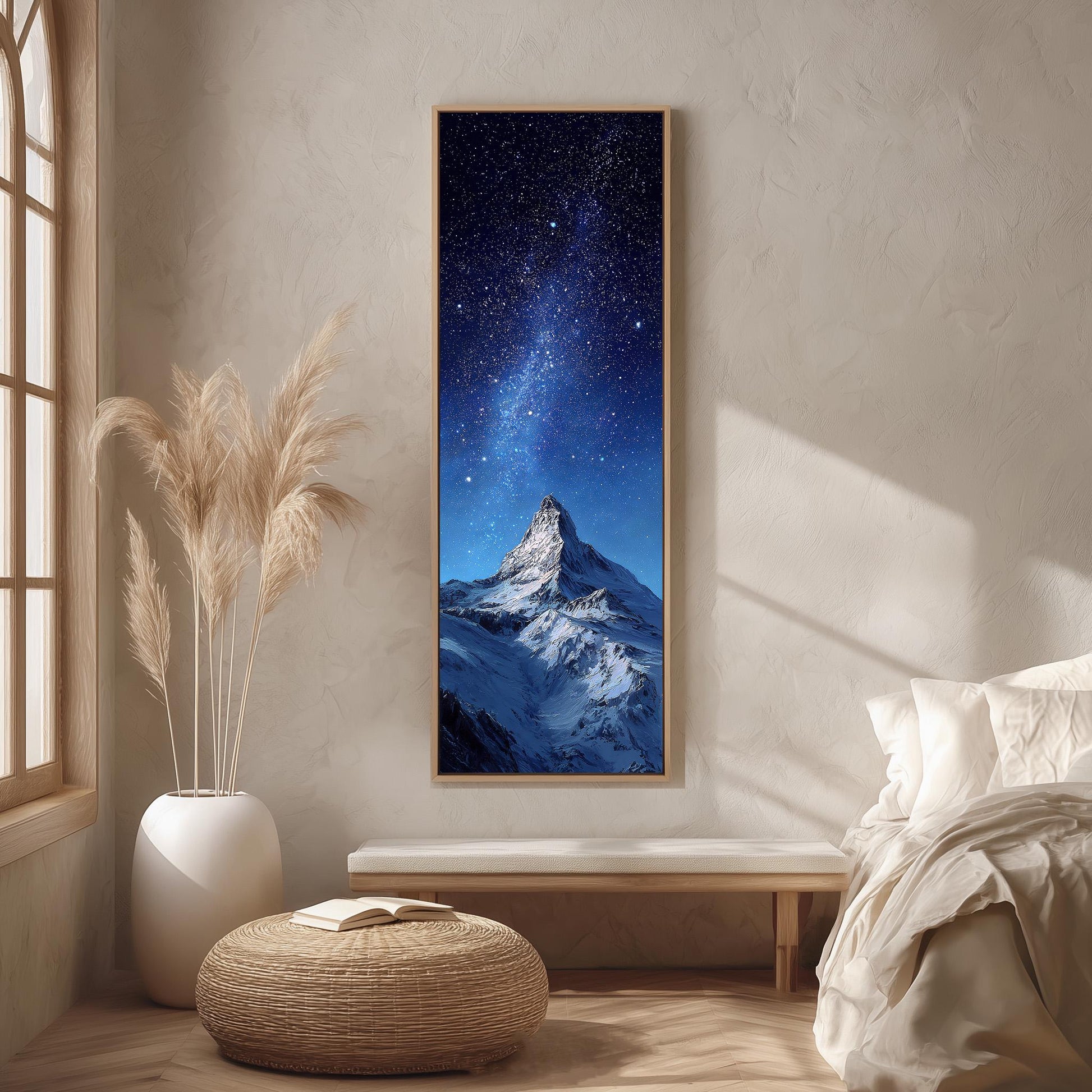 a serene bedroom setting with a large framed picture of a snowy mountain peak under a starry night sky hanging on the wall. The room has a white bed, a wicker ottoman, and a potted plant, creating a cozy and inviting atmosphere.