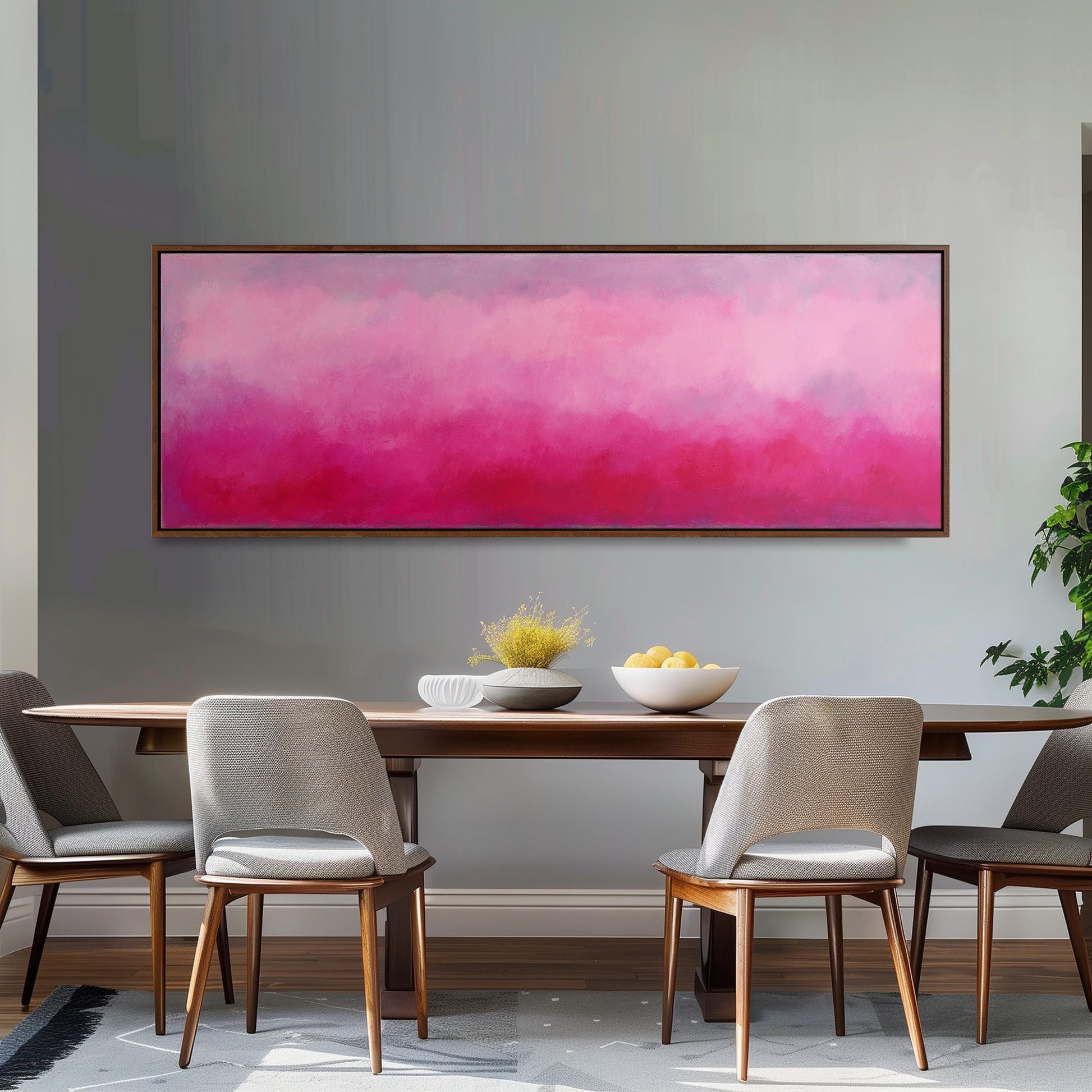 a dining room with a wooden table and four chairs. Above the table, there is a large painting with a pink and purple gradient background. On the table, there is a bowl of fruit and a vase of flowers.