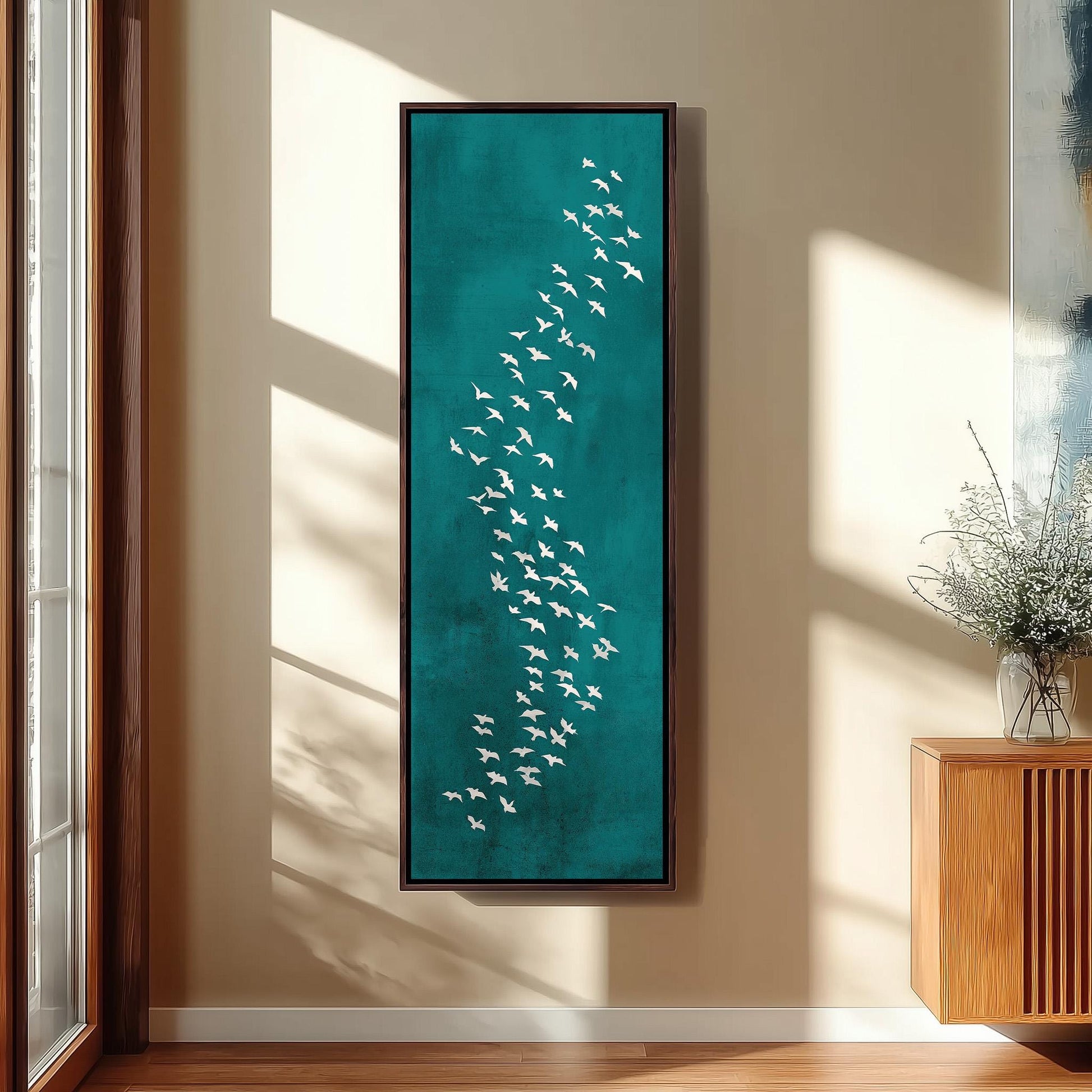 a large framed artwork depicting a flock of white birds on a teal background, hanging on a wall in a room with a wooden cabinet and a vase of flowers.