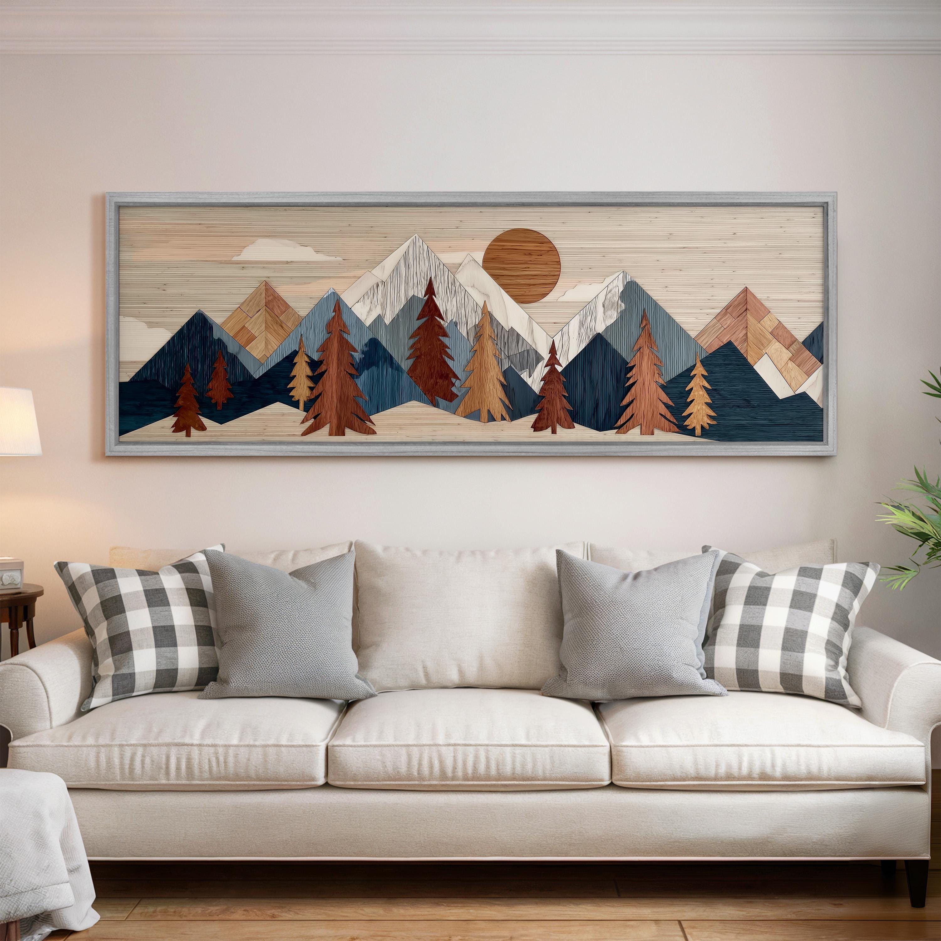 a large wooden framed artwork depicting a mountain landscape with pine trees and a sun in the sky, hanging on a wall above a white couch with pillows.