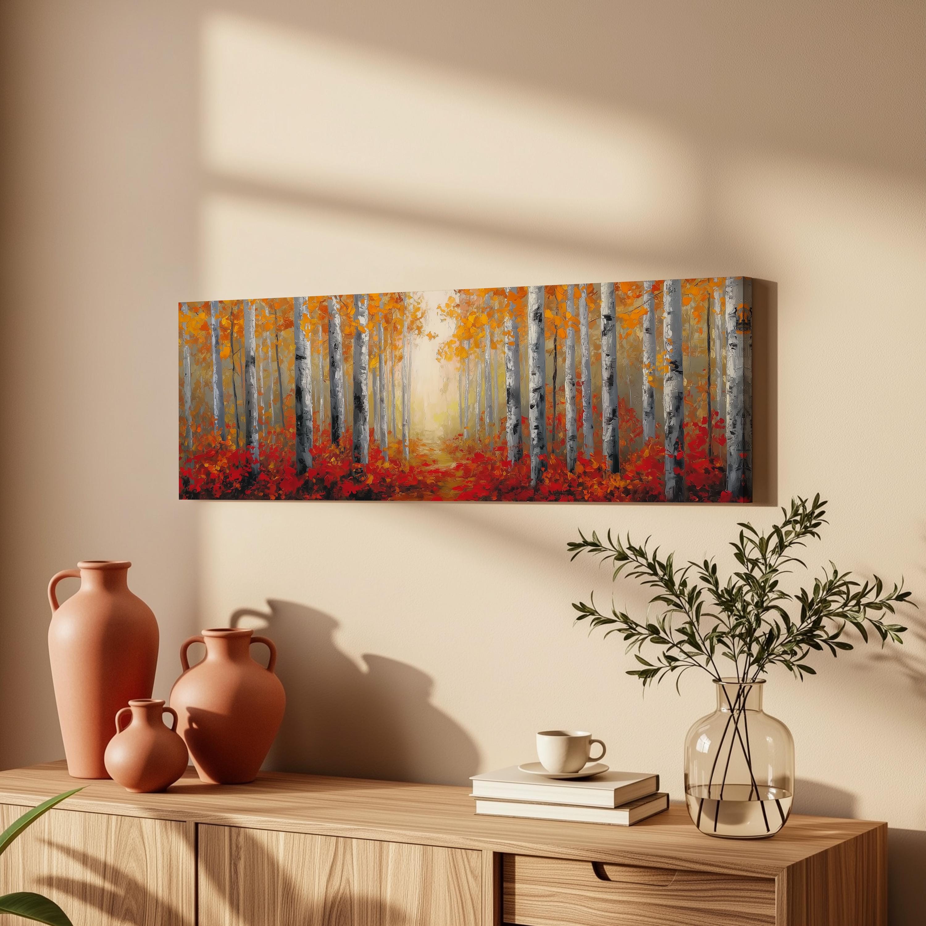 a wooden cabinet with a painting of a forest scene hanging on the wall above it. The painting depicts a path leading through a forest of tall trees with vibrant orange and red foliage. The cabinet also has three orange vases, a white coffee cup, and a stack of books on top.