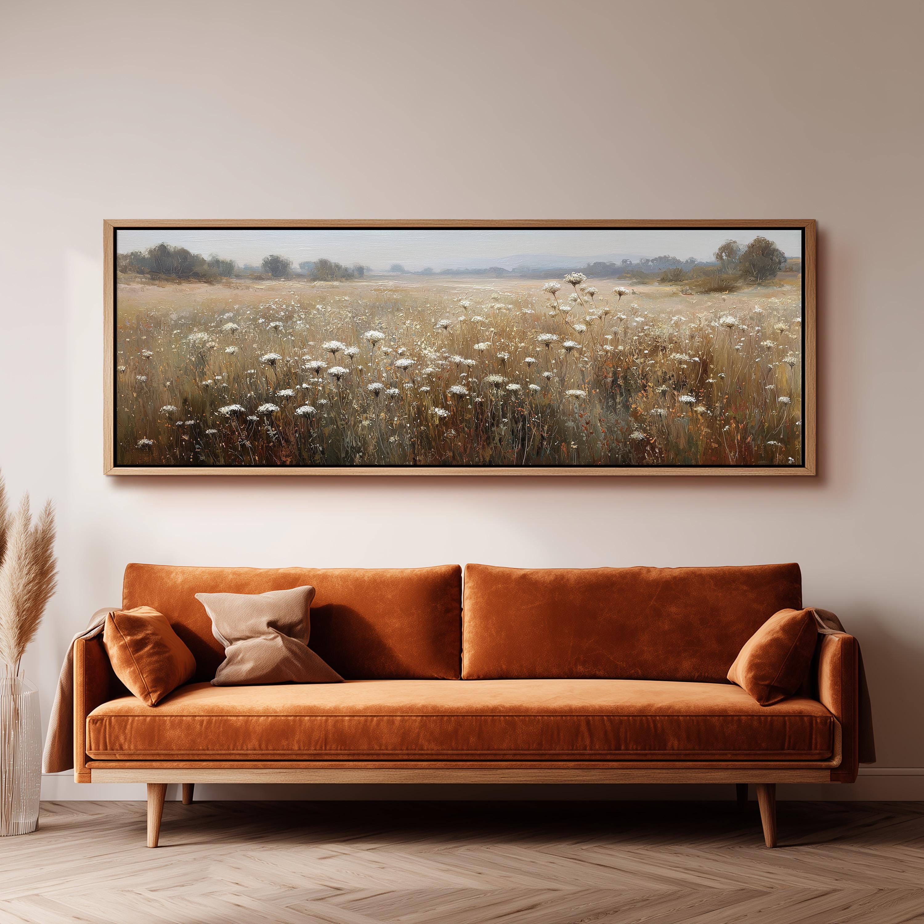 a large, framed painting of a field with tall grass and wildflowers hanging on a wall above a brown sofa with orange cushions.