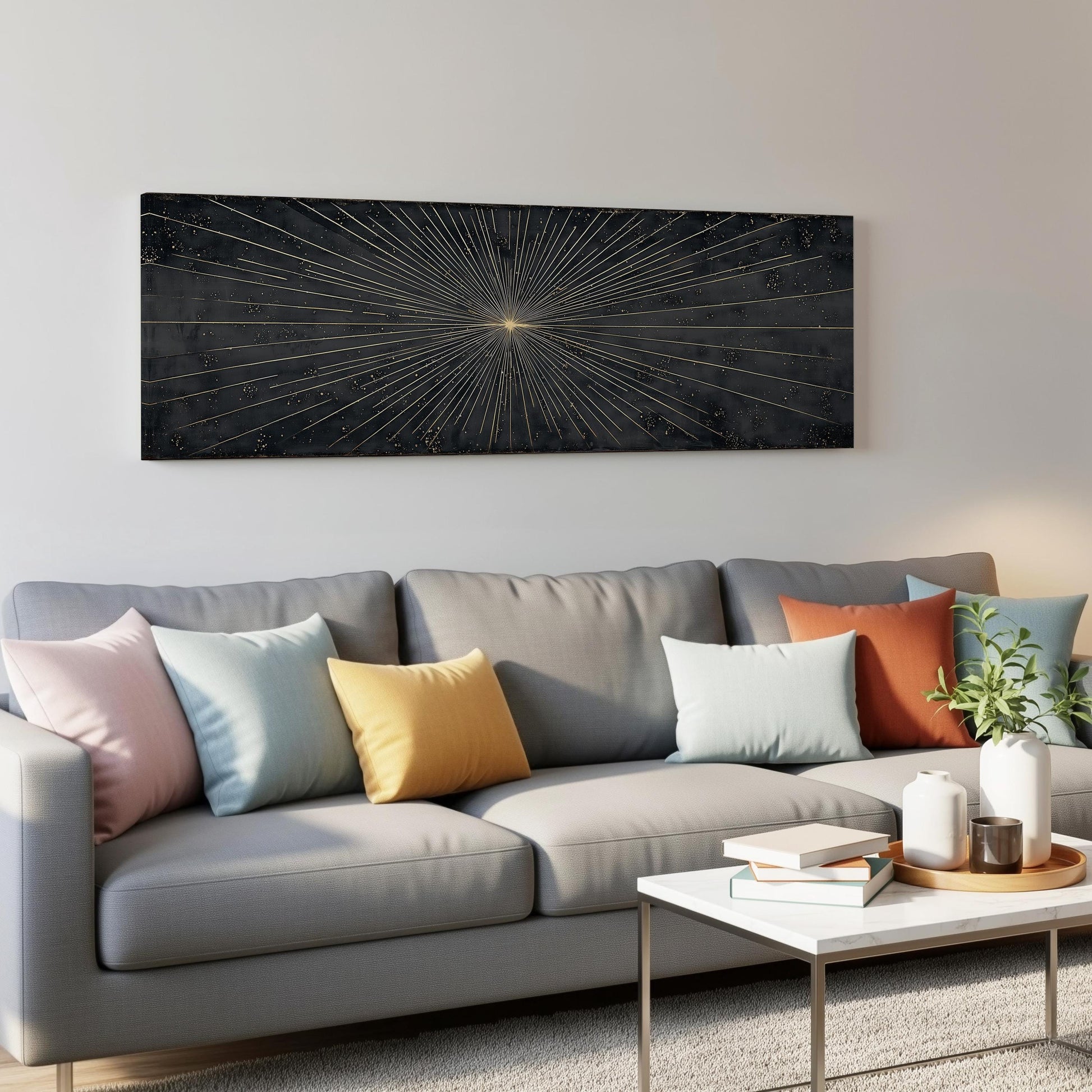 a modern living room with a gray sofa adorned with colorful throw pillows. Above the sofa, a large black and gold abstract painting hangs on the wall, adding a striking visual element to the space. In front of the sofa, there is a white coffee table with a stack of books and a small plant, creating a cozy and inviting atmosphere.