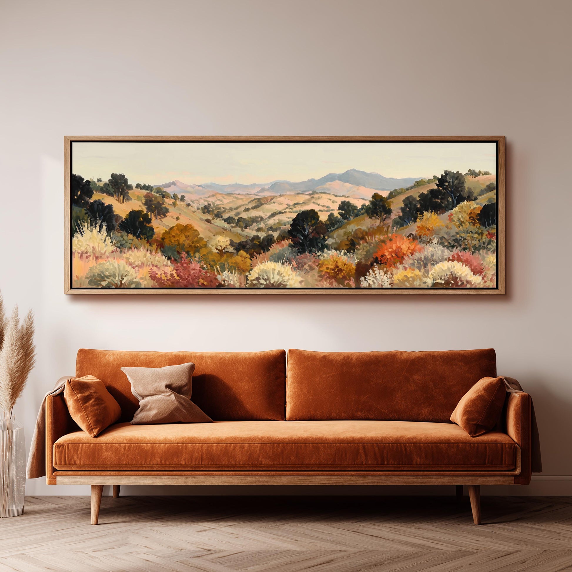 a large, framed painting of a mountainous landscape hanging on a wall above a brown sofa with orange cushions.