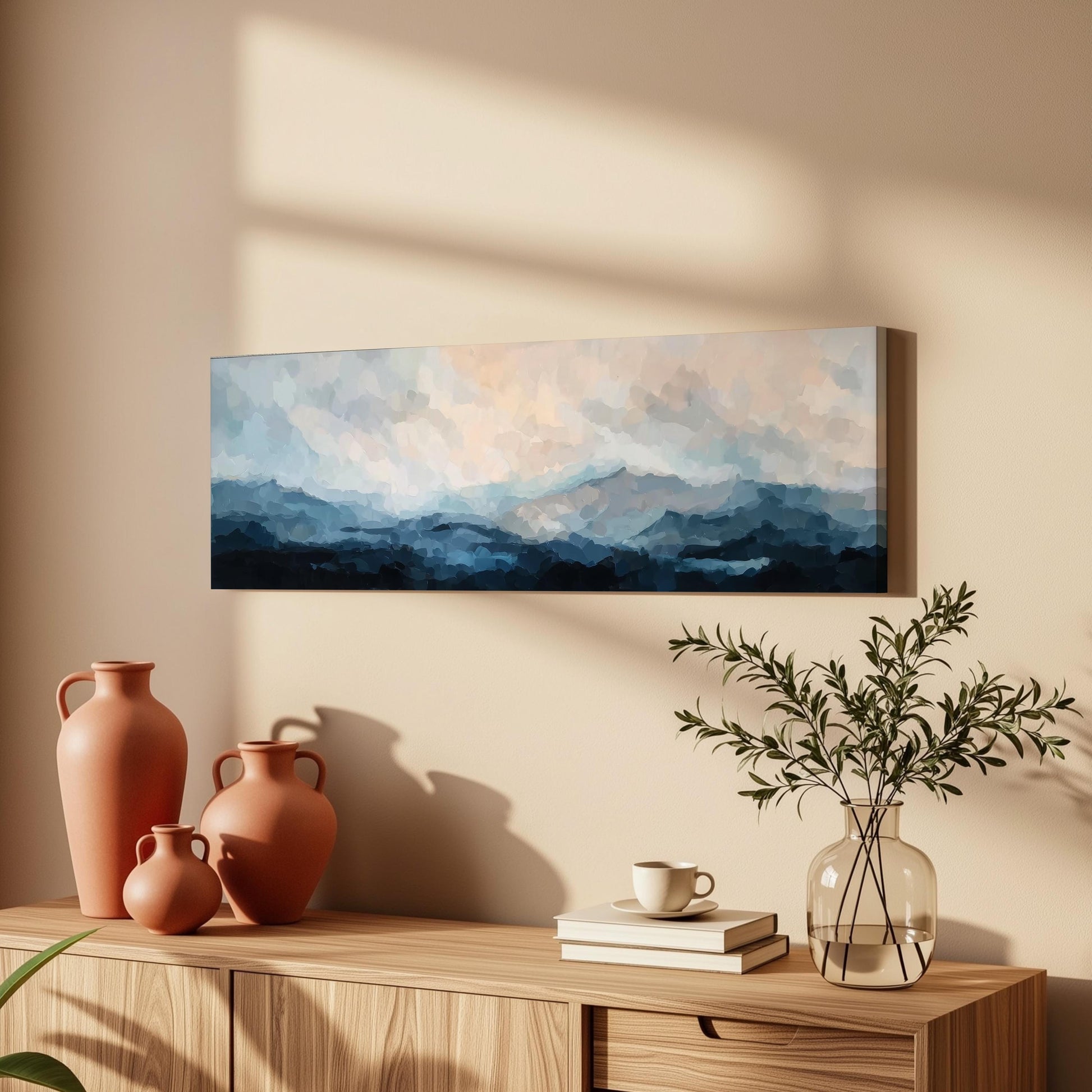 a wooden cabinet with a painting of a mountain landscape hanging on the wall above it. The cabinet also has three orange vases and a glass vase with greenery, as well as a stack of books and a coffee cup on the surface.