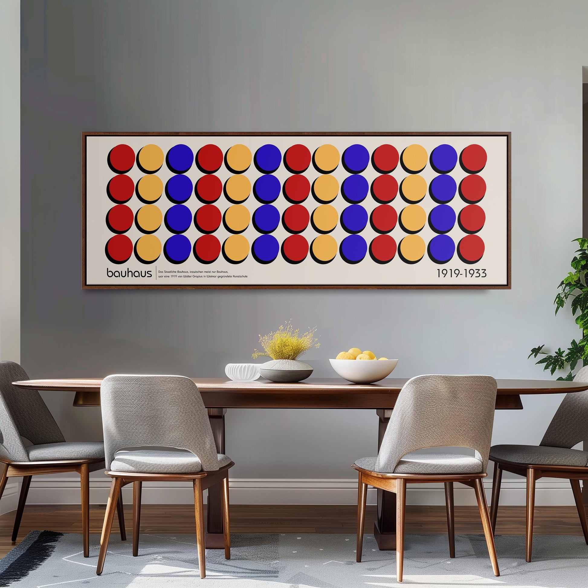 a dining room with a wooden table and four chairs. Above the table, there is a large framed poster with a repeating pattern of circles in various colors. The poster is hanging on a gray wall, and the dining table is set with a bowl of fruit and a vase.