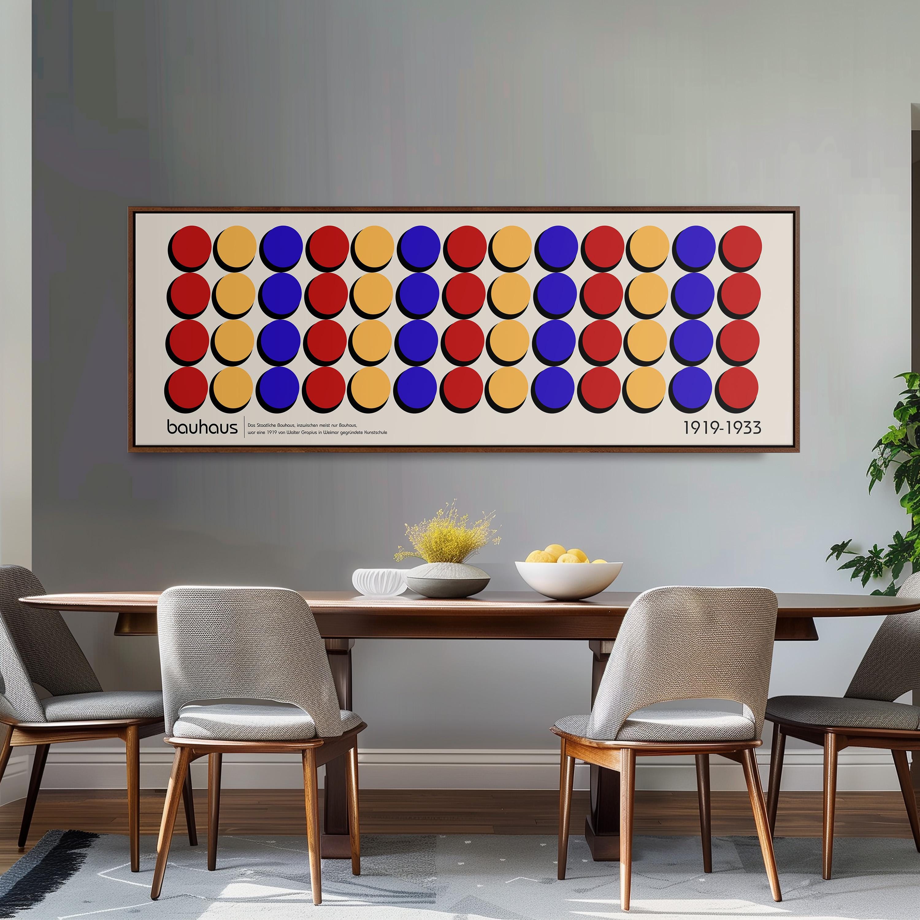 a dining room with a wooden table and four chairs. Above the table, there is a large framed poster with a repeating pattern of circles in various colors. The poster is hanging on a gray wall, and the dining table is set with a bowl of fruit and a vase.