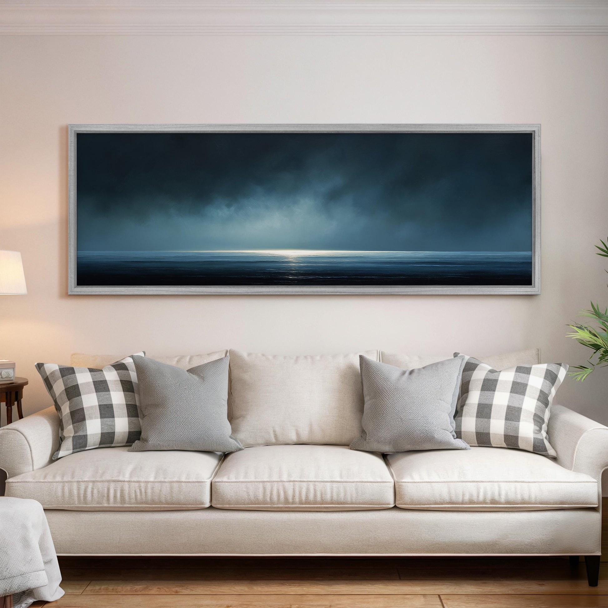 a large, framed painting of a serene ocean scene hanging on a wall above a white sofa with gray and white patterned throw pillows.