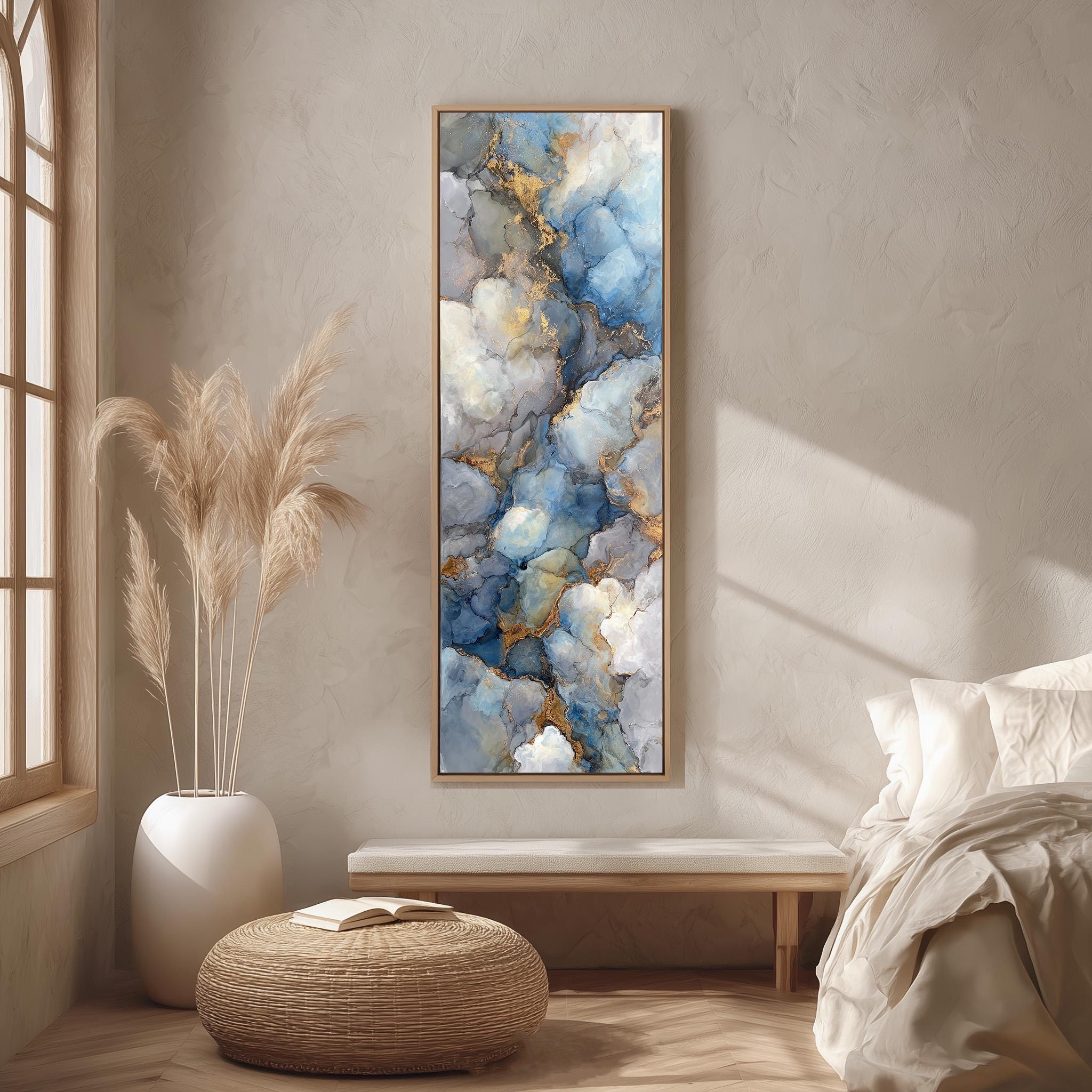 a large abstract painting hanging on a wall in a bedroom setting. The painting has a blue and white color scheme with a textured, layered appearance. The room includes a bed, a wicker ottoman, and a potted plant, creating a cozy and inviting atmosphere.