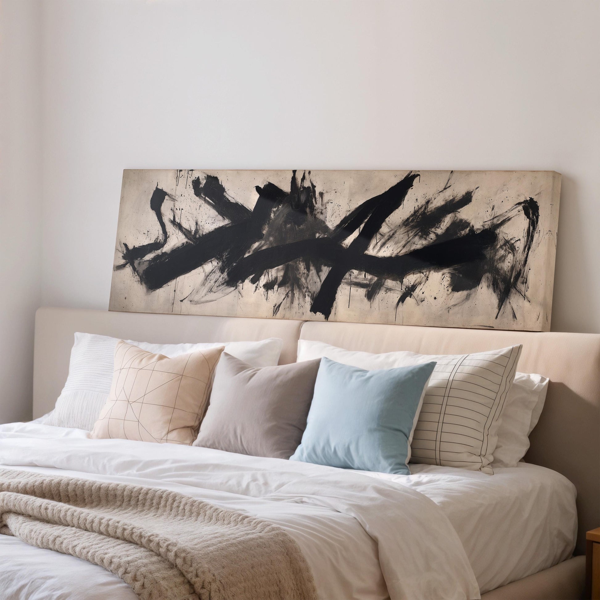 a large bed with a white comforter and a variety of pillows, including a blue pillow and a striped pillow. Above the bed, there is a large abstract painting with bold black lines and splatters.