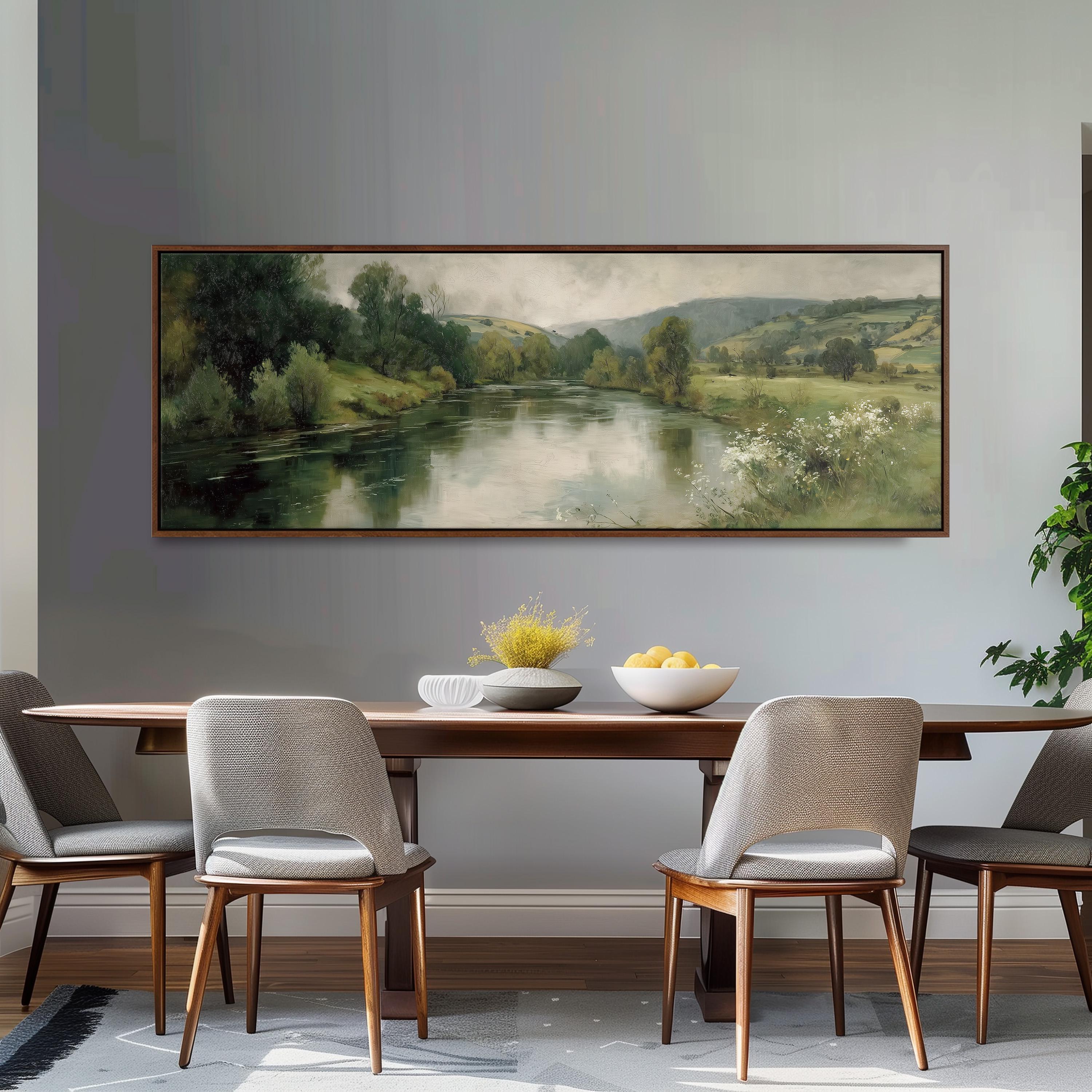 a large painting of a serene landscape hanging on a wall above a wooden dining table with four chairs. The painting depicts a peaceful countryside scene with a river, trees, and rolling hills. The table is set with a bowl of fruit and a vase of flowers, creating a warm and inviting atmosphere.