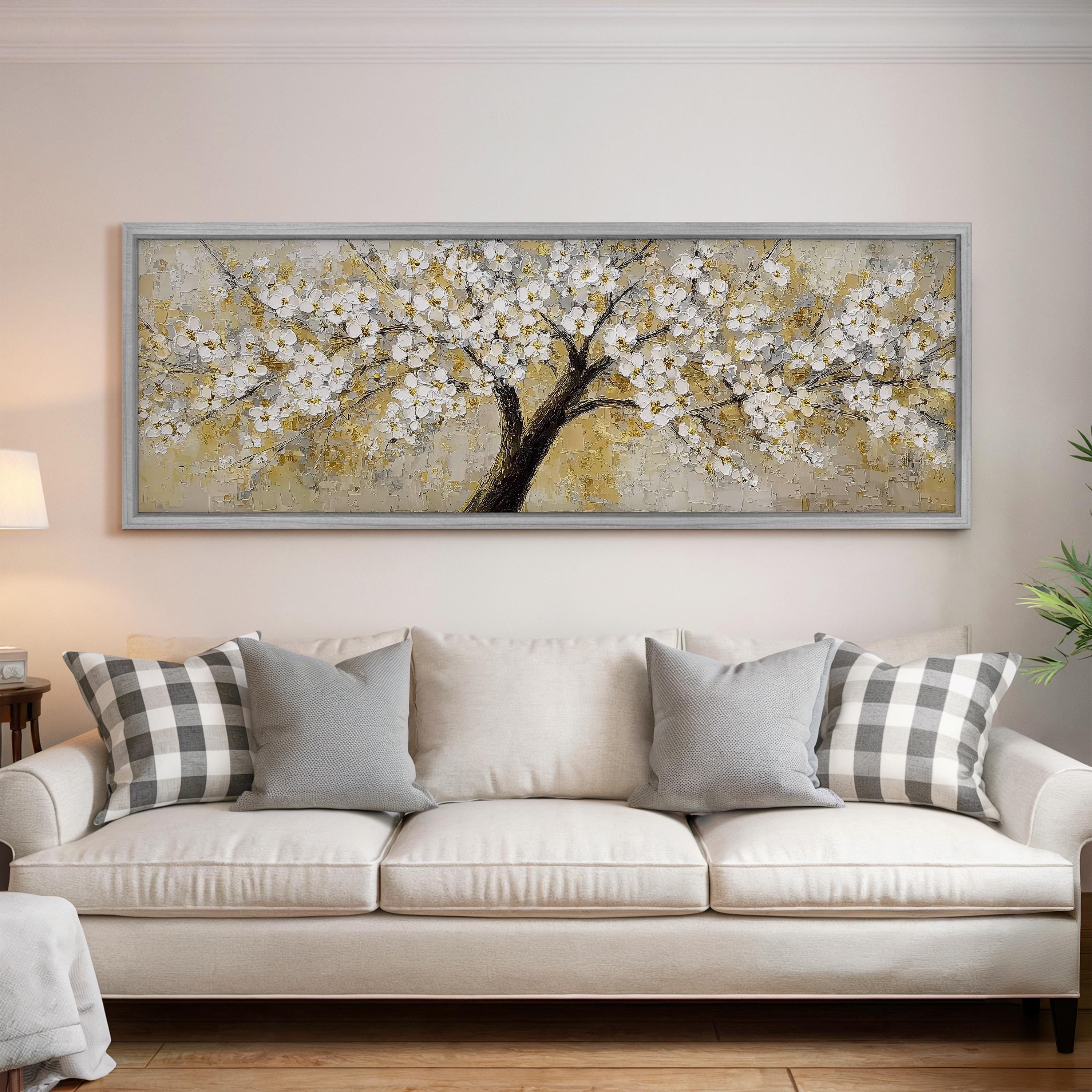 a large, framed painting of a tree with white flowers hanging above a white couch with gray and white pillows.