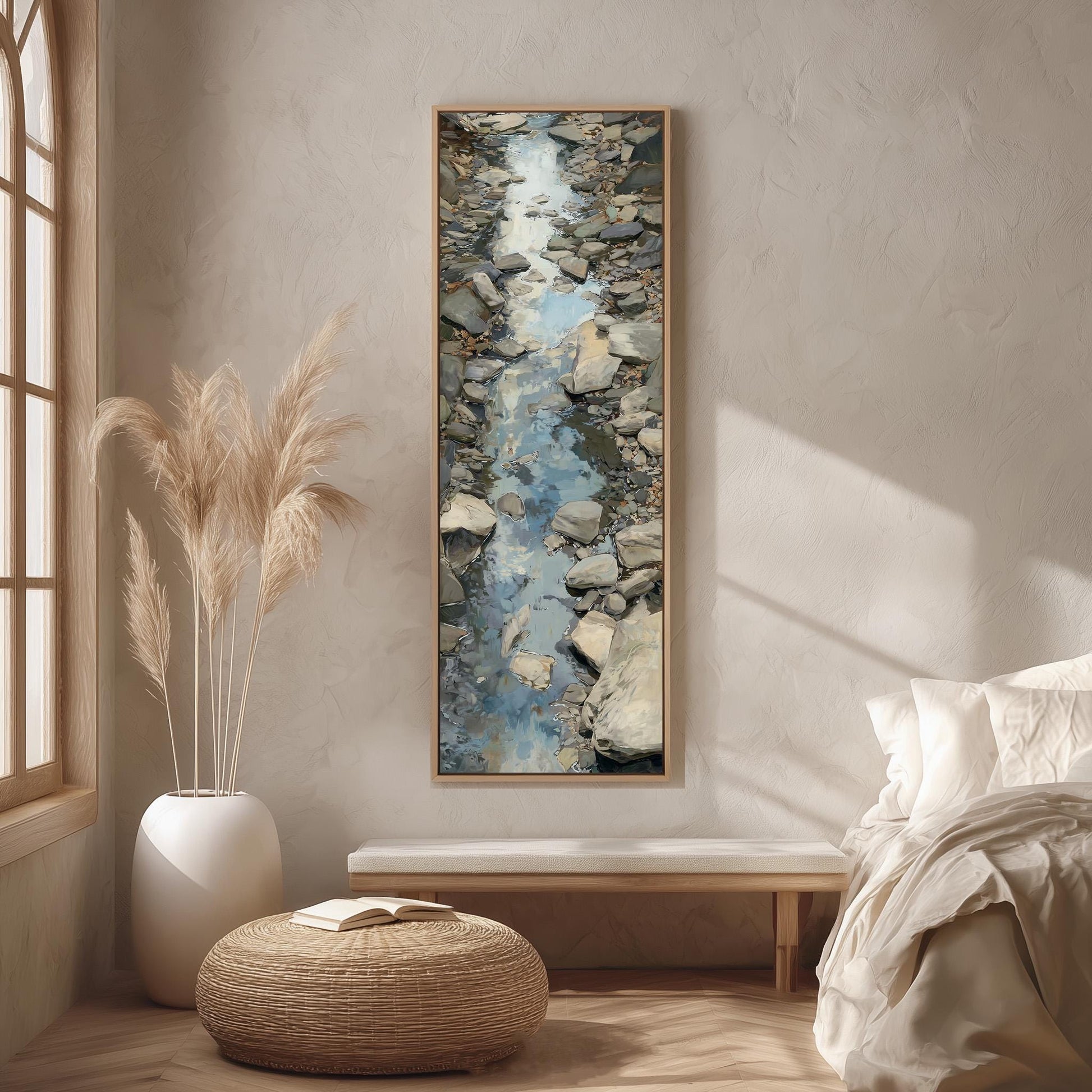 a serene bedroom scene with a large painting of a rocky stream hanging on the wall. The painting is framed in a wooden frame and is the focal point of the room. The room features a bed with white linens, a wicker ottoman, and a potted plant. The overall color scheme of the room is neutral, with the painting adding a touch of natural beauty to the space.