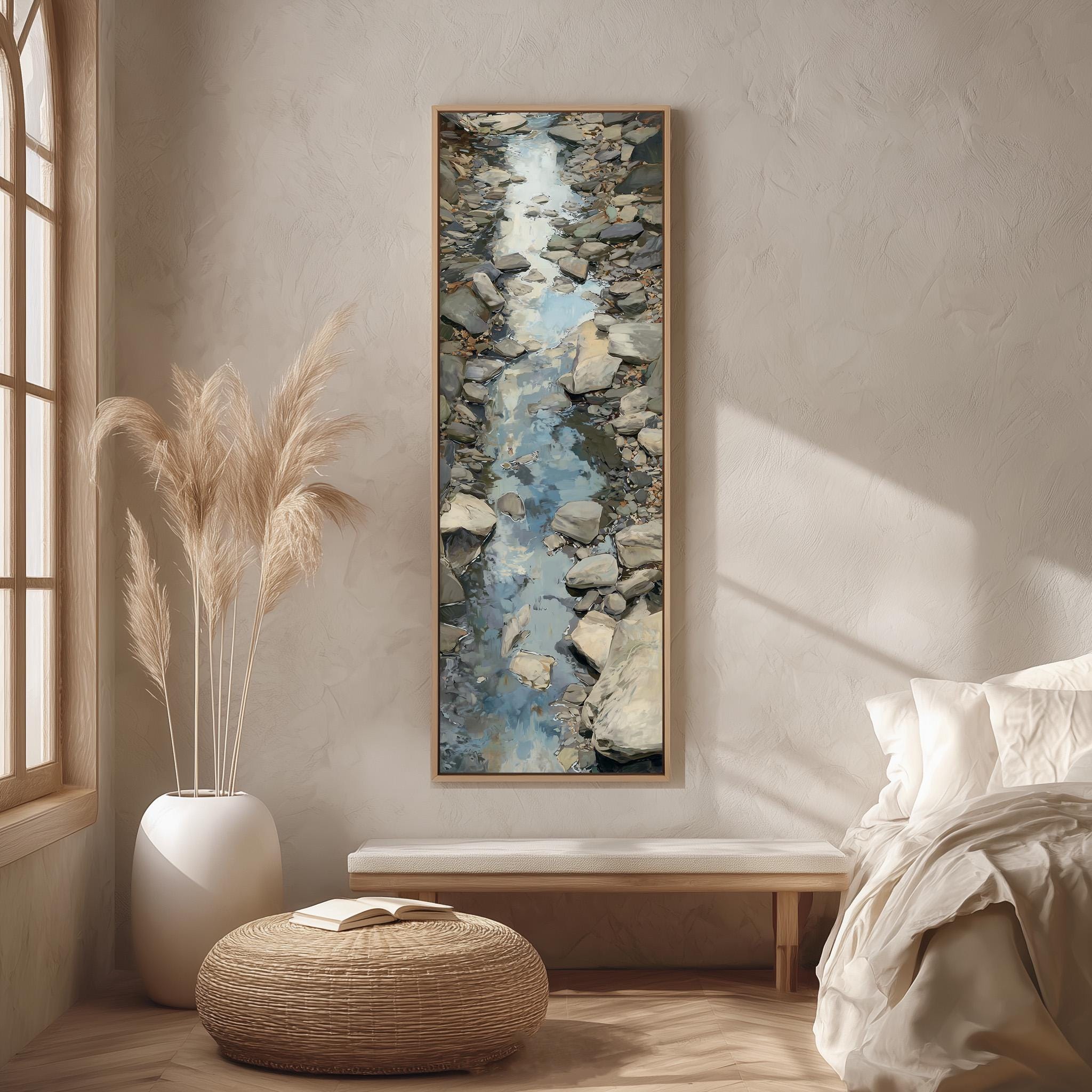 a serene bedroom scene with a large painting of a rocky stream hanging on the wall. The painting is framed in a wooden frame and is the focal point of the room. The room features a bed with white linens, a wicker ottoman, and a potted plant. The overall color scheme of the room is neutral, with the painting adding a touch of natural beauty to the space.