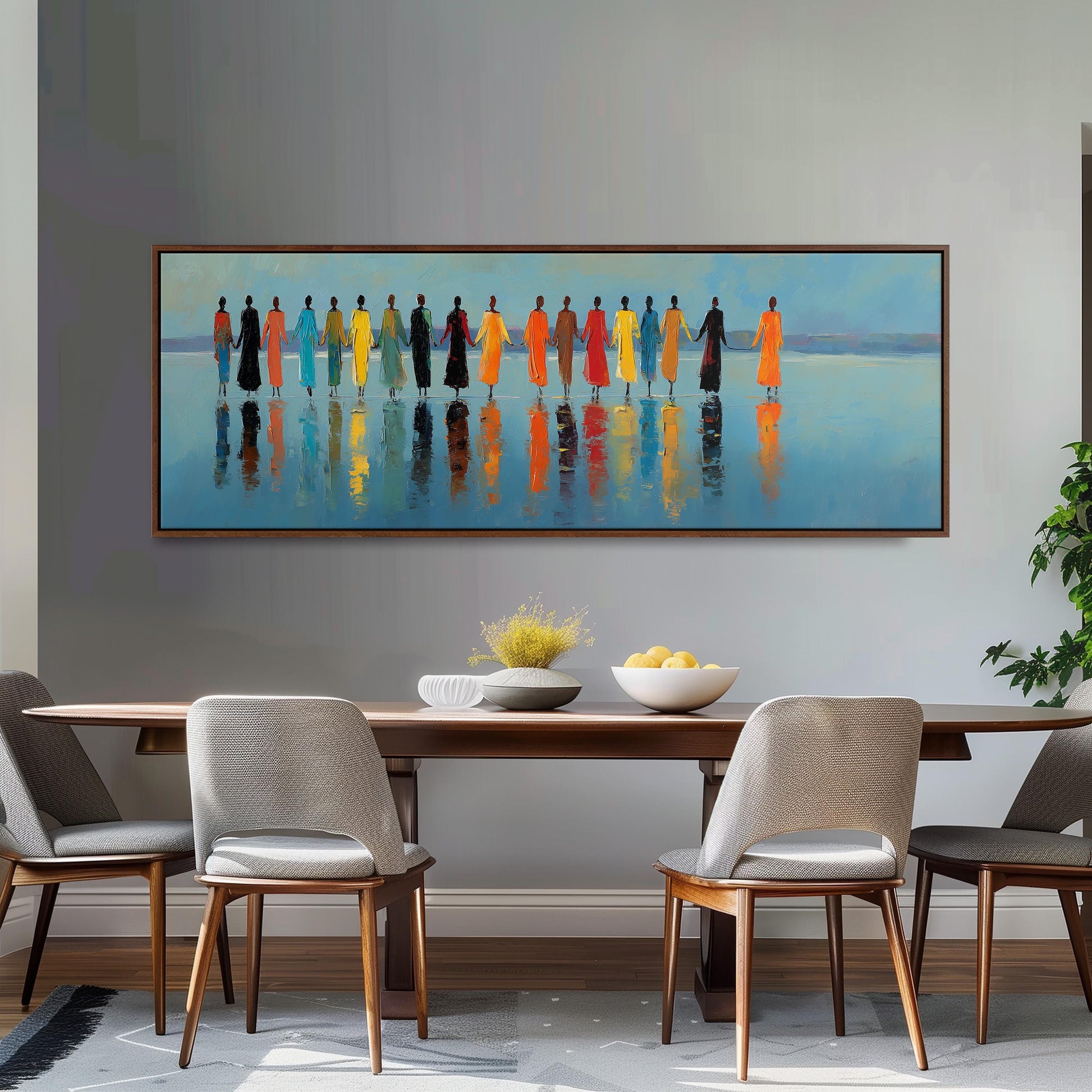 a large painting of a group of people standing in a line, with their reflections visible in the water below. The painting is hung on a wall above a wooden dining table with four chairs. The table is set with a bowl of fruit and a vase, creating a cozy and inviting atmosphere.