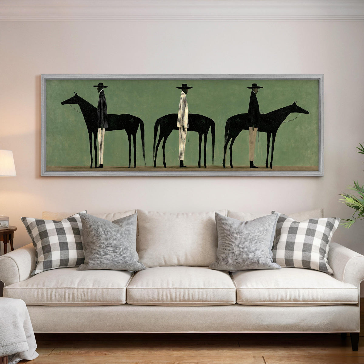 Three stylized cowboys with horses on sage green panoramic canvas print above a white sofa in a bright living room