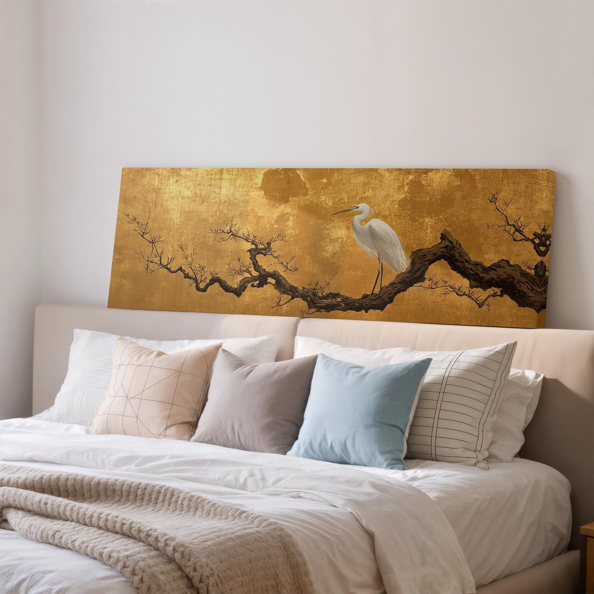 a large bed with a white comforter and several pillows, including a blue pillow and a striped pillow. Above the bed, there is a gold-colored painting of a bird perched on a branch.