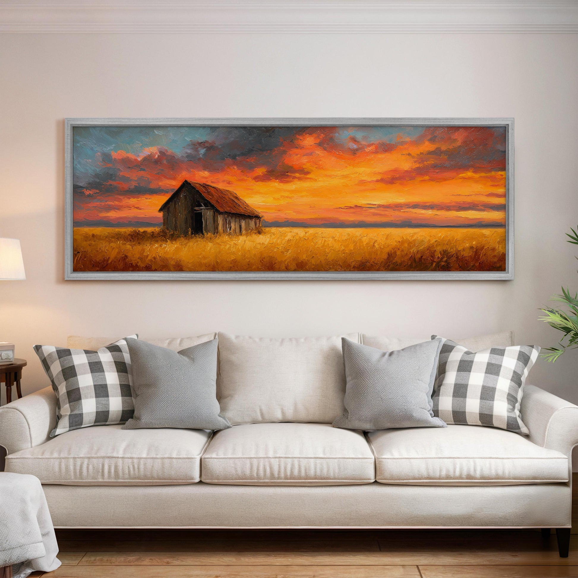 a large painting of a barn and a field with a colorful sunset sky above it, hanging on a wall above a white couch with patterned throw pillows.