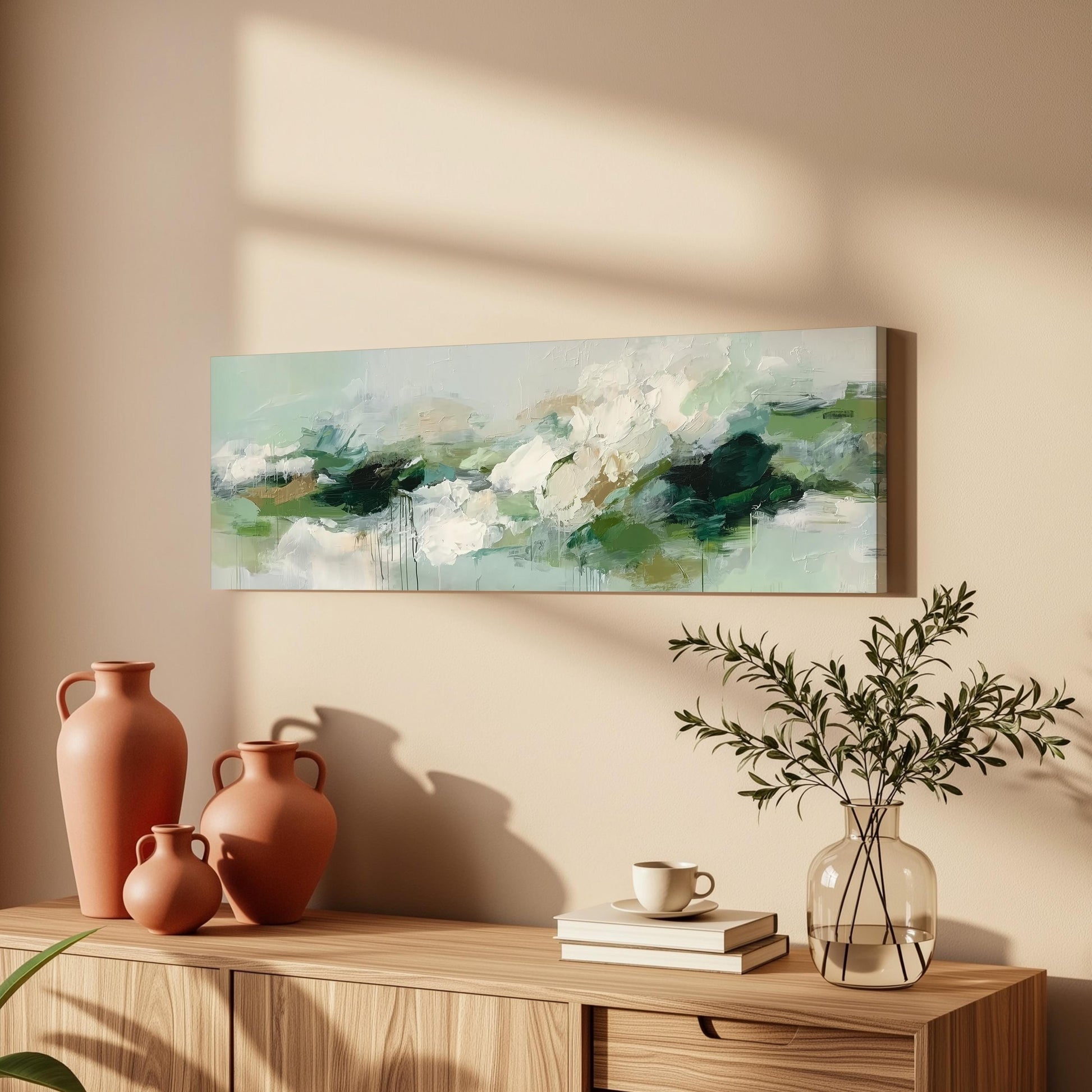 a wooden cabinet with a painting hanging on the wall above it, along with three orange vases and a glass vase with greenery. The cabinet also has a stack of books and a coffee cup on top.