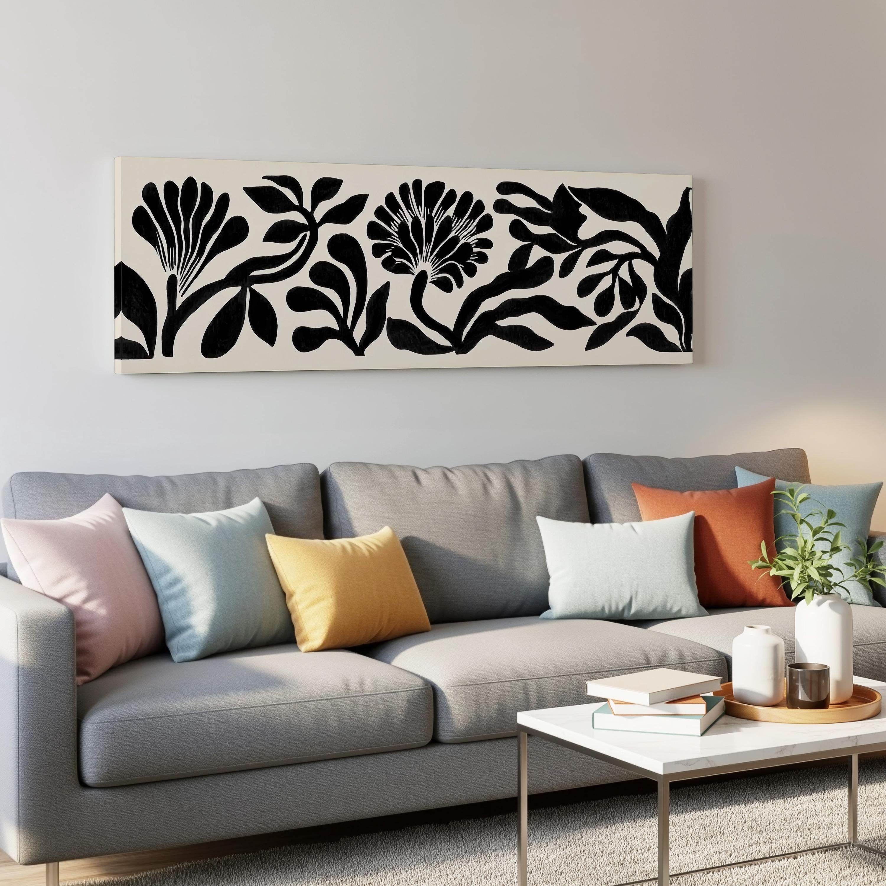 a modern living room with a gray sofa adorned with colorful throw pillows. Above the sofa, there is a large black and white floral artwork hanging on the wall. In front of the sofa, there is a white coffee table with a stack of books and a small plant.