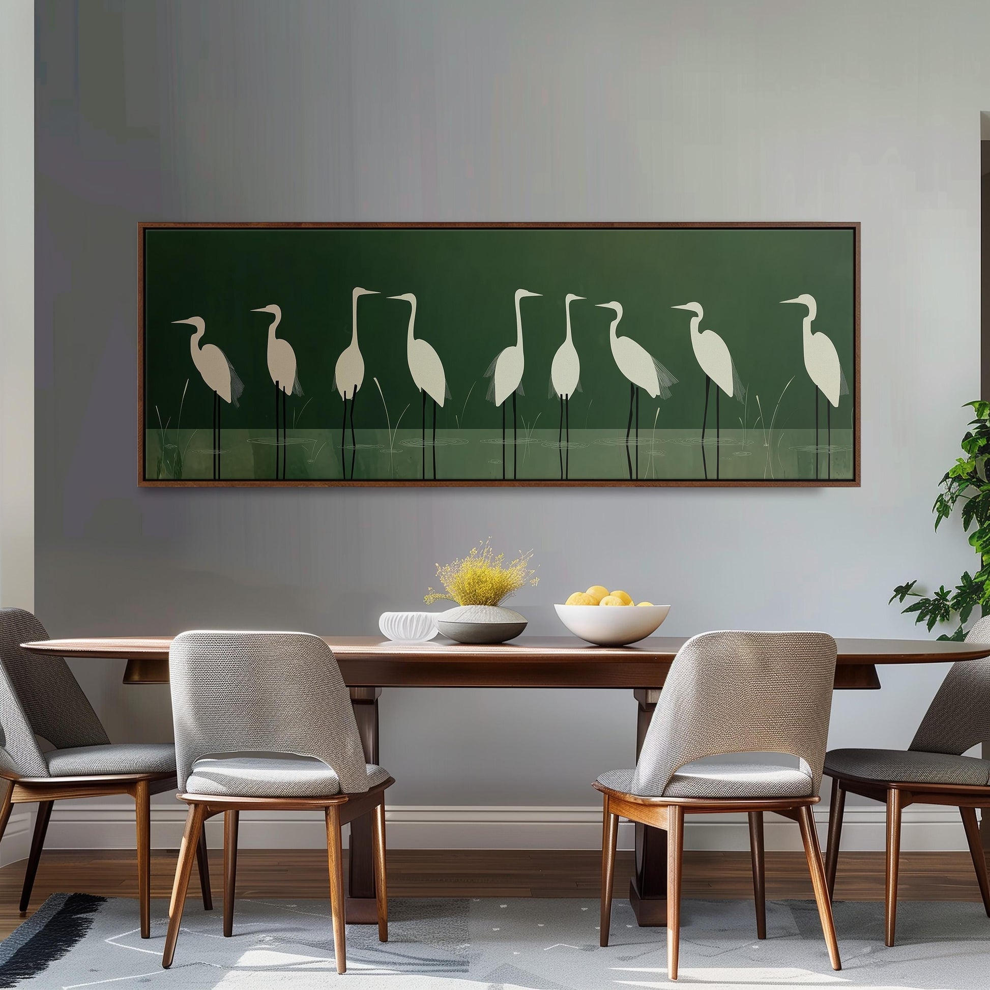 a dining room with a wooden table and four chairs. Above the table, there is a large framed picture of a group of white birds, possibly herons, on a green background. The table is set with a bowl of fruit and a vase of flowers, creating a cozy and inviting atmosphere.