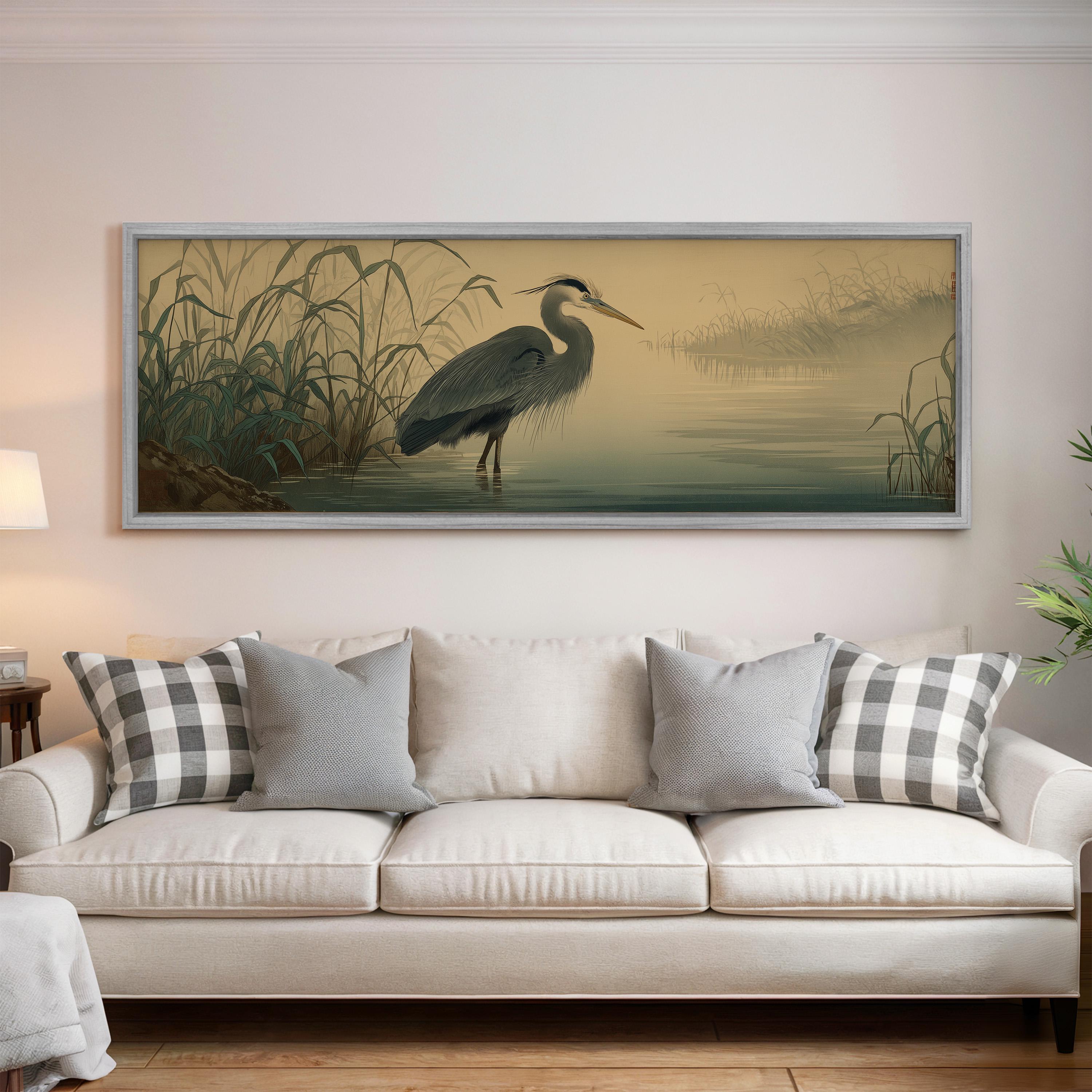 a large framed painting of a heron standing in a body of water, hanging on a wall above a white sofa with decorative pillows.