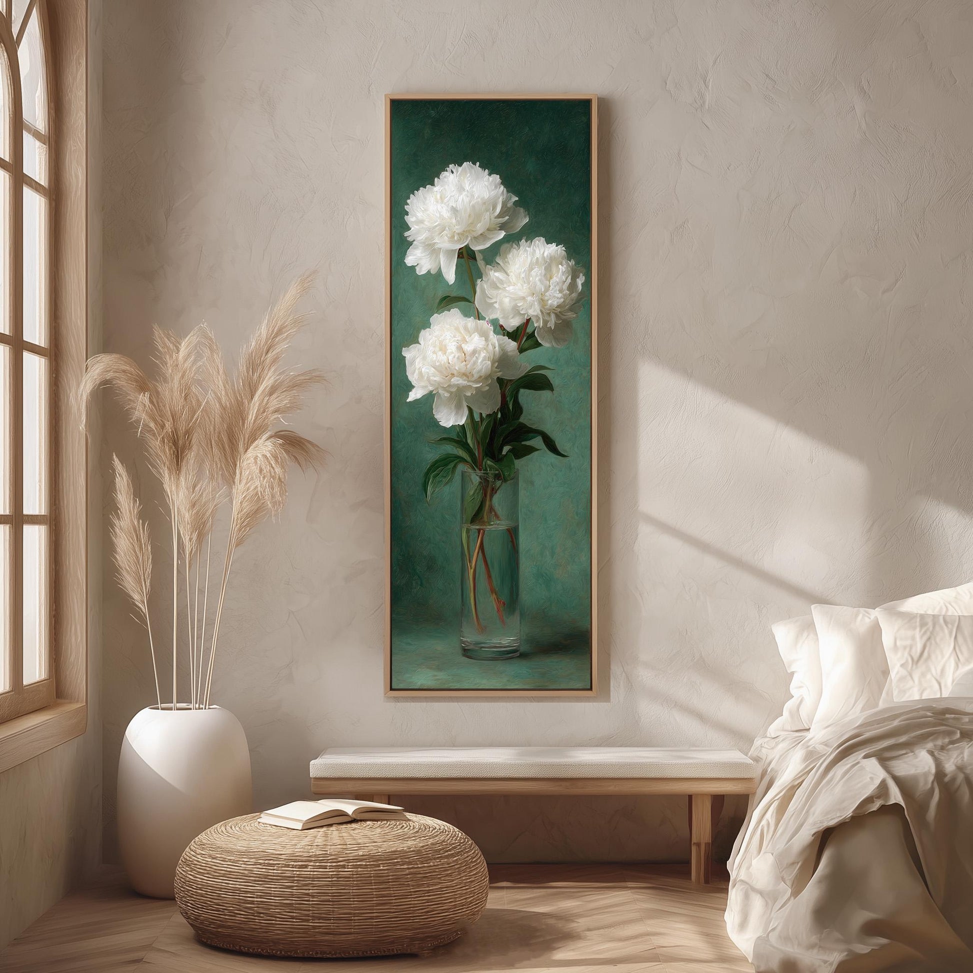 a serene and elegant living room with a large painting of white peonies hanging on the wall. The painting is framed in a wooden frame and is placed above a white bench. To the left of the painting, there is a tall white vase filled with long, light-colored peonies. The room also includes a bed with white bedding and a woven ottoman in the foreground.