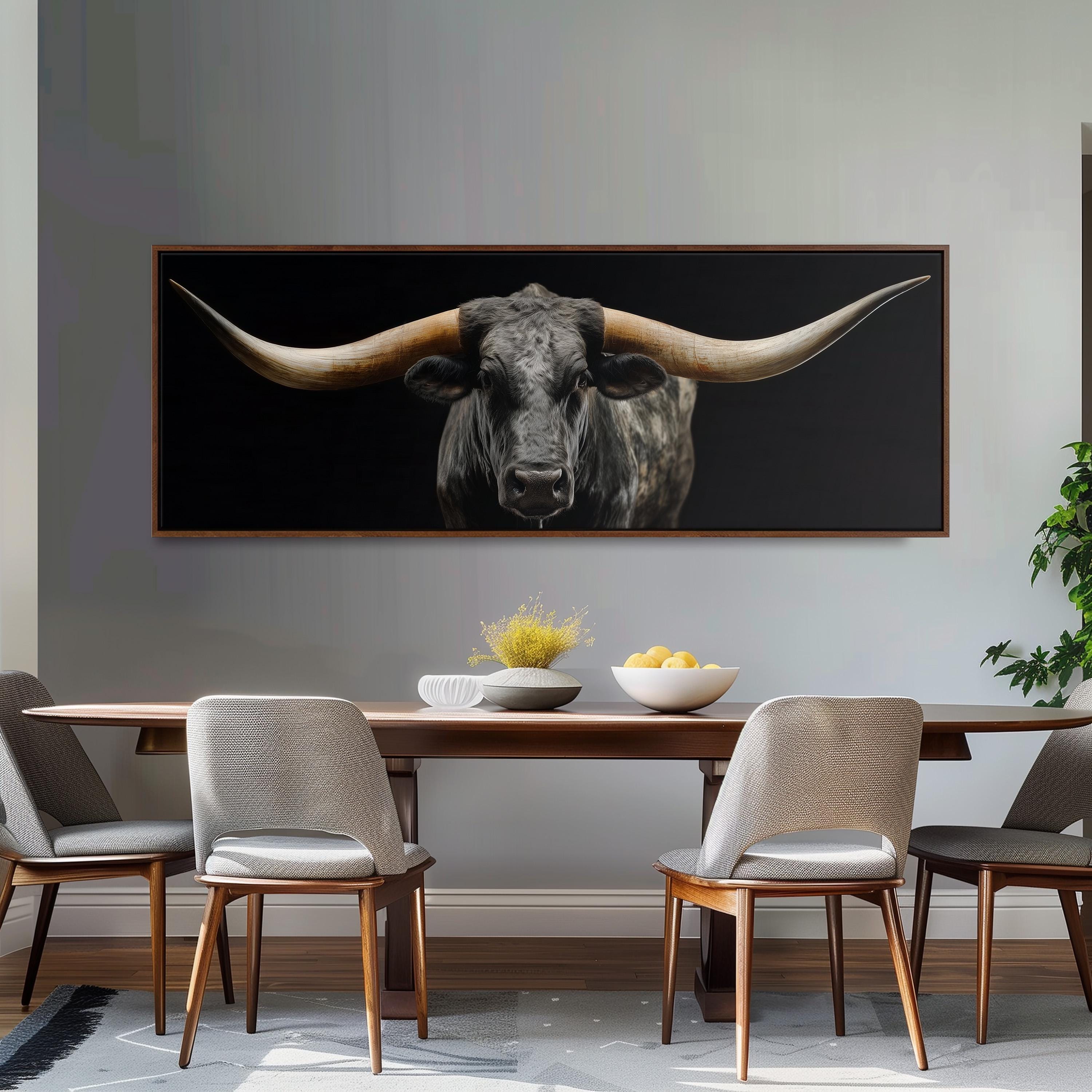 a large framed picture of a bull with long horns hanging on a wall above a wooden dining table with four chairs.