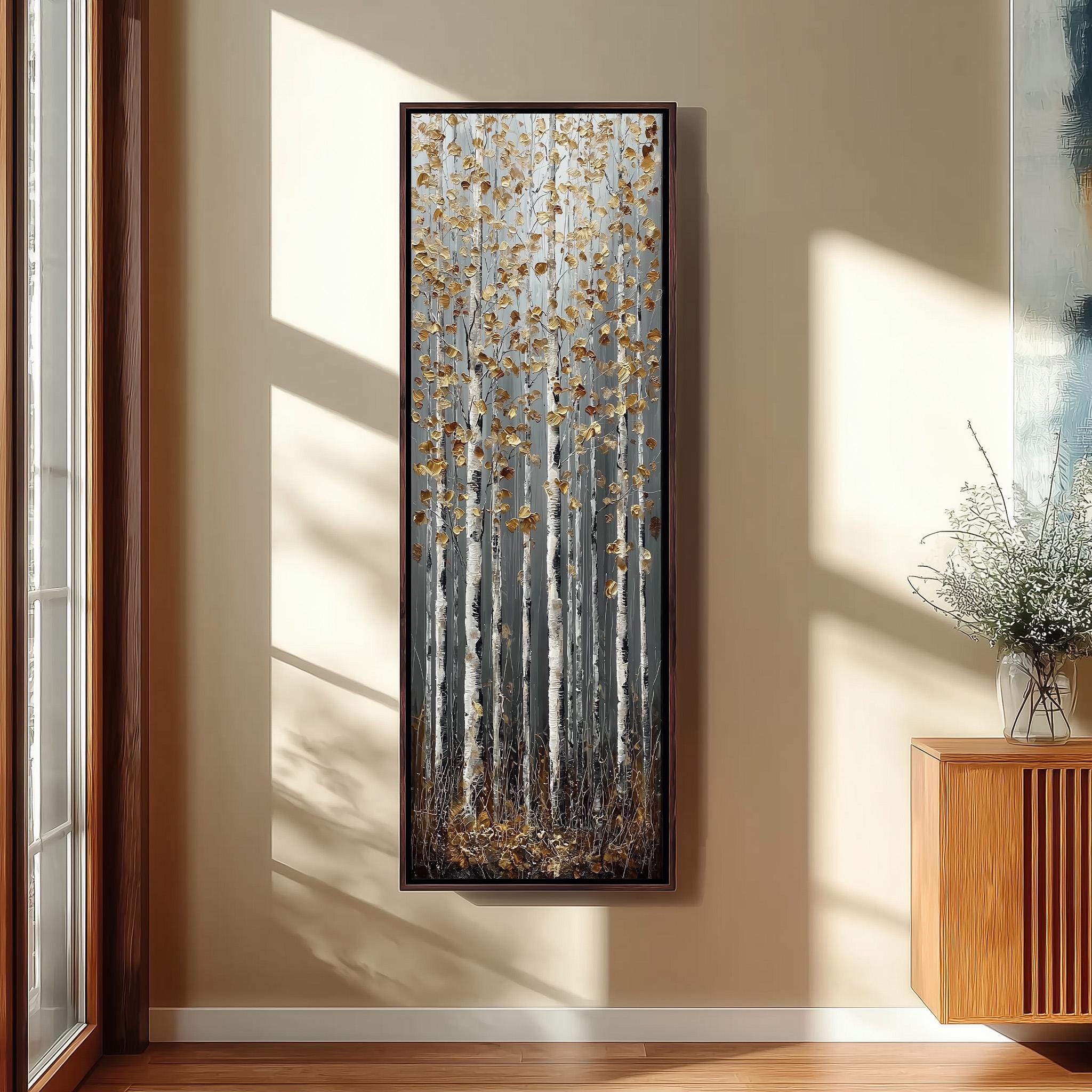 a large framed painting of a forest scene with tall trees and golden leaves, hanging on a wall in a room with a wooden cabinet and a vase of flowers.