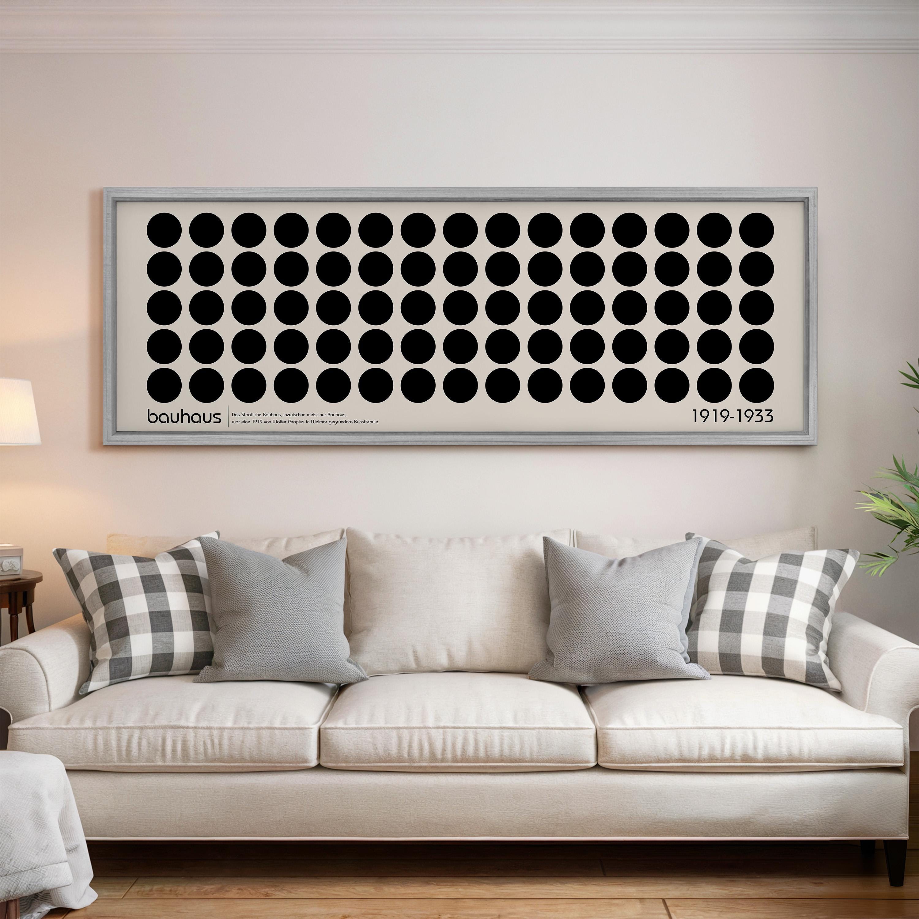 a large framed artwork with a repeating pattern of black circles on a white background, hanging above a white sofa with gray and white plaid throw pillows.