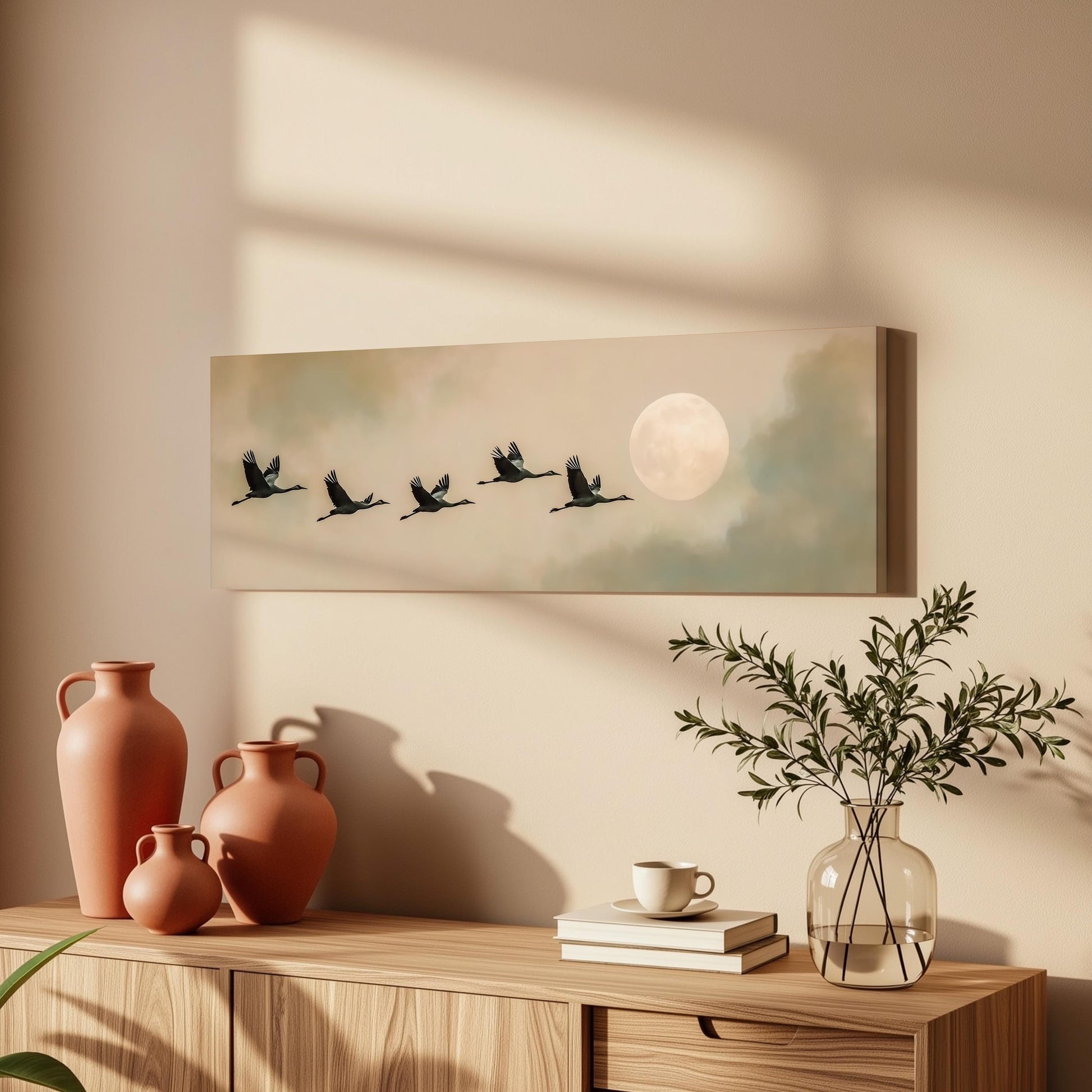 a wooden cabinet with a vase, a coffee cup, and a stack of books on top. Above the cabinet, there is a framed painting depicting a flock of birds flying in formation, with a full moon in the background.