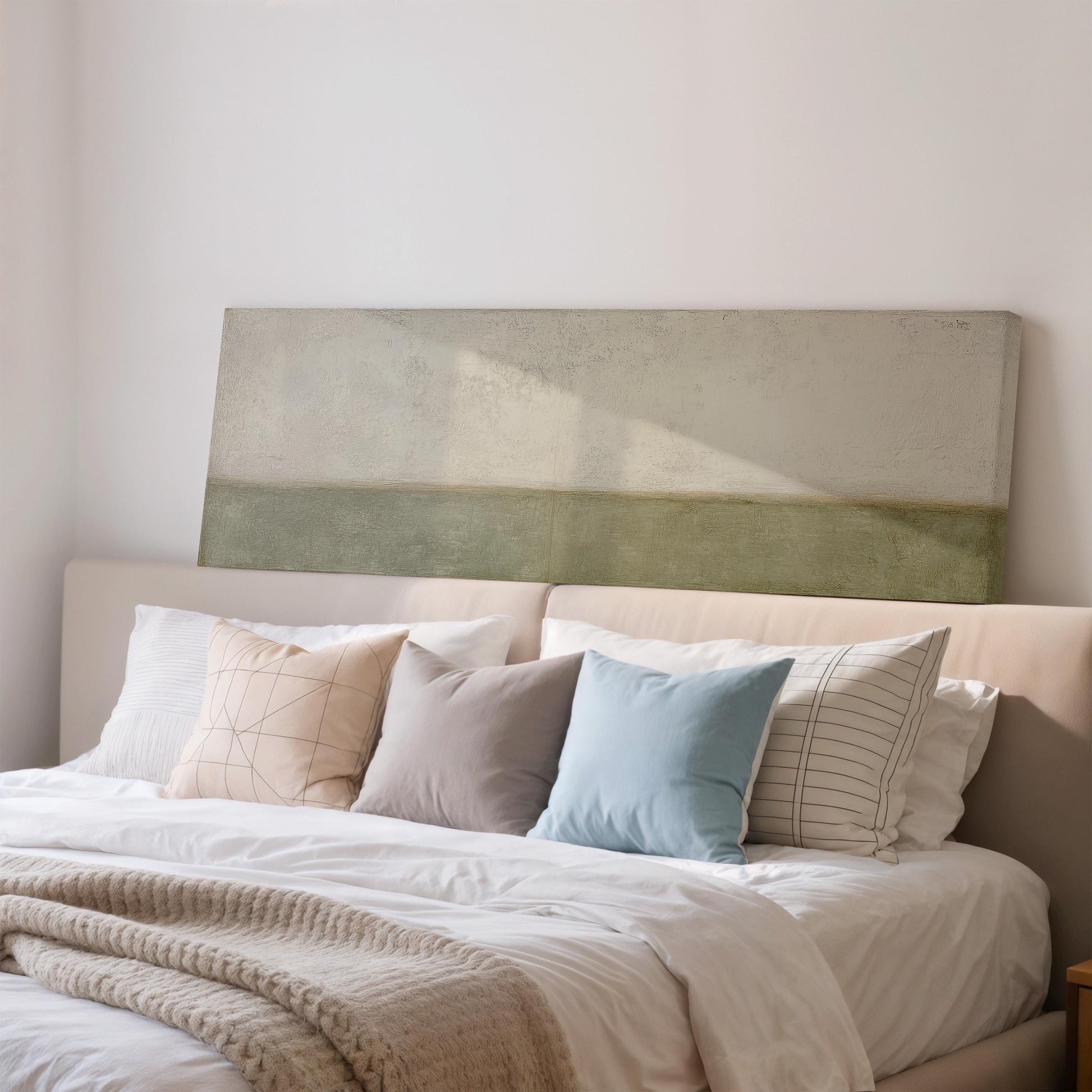 a neatly made bed with a white comforter and a variety of colorful pillows. The pillows include a blue one, a gray one, and a striped one. The bed is positioned against a wall, and there is a large piece of artwork or painting hanging above it.
