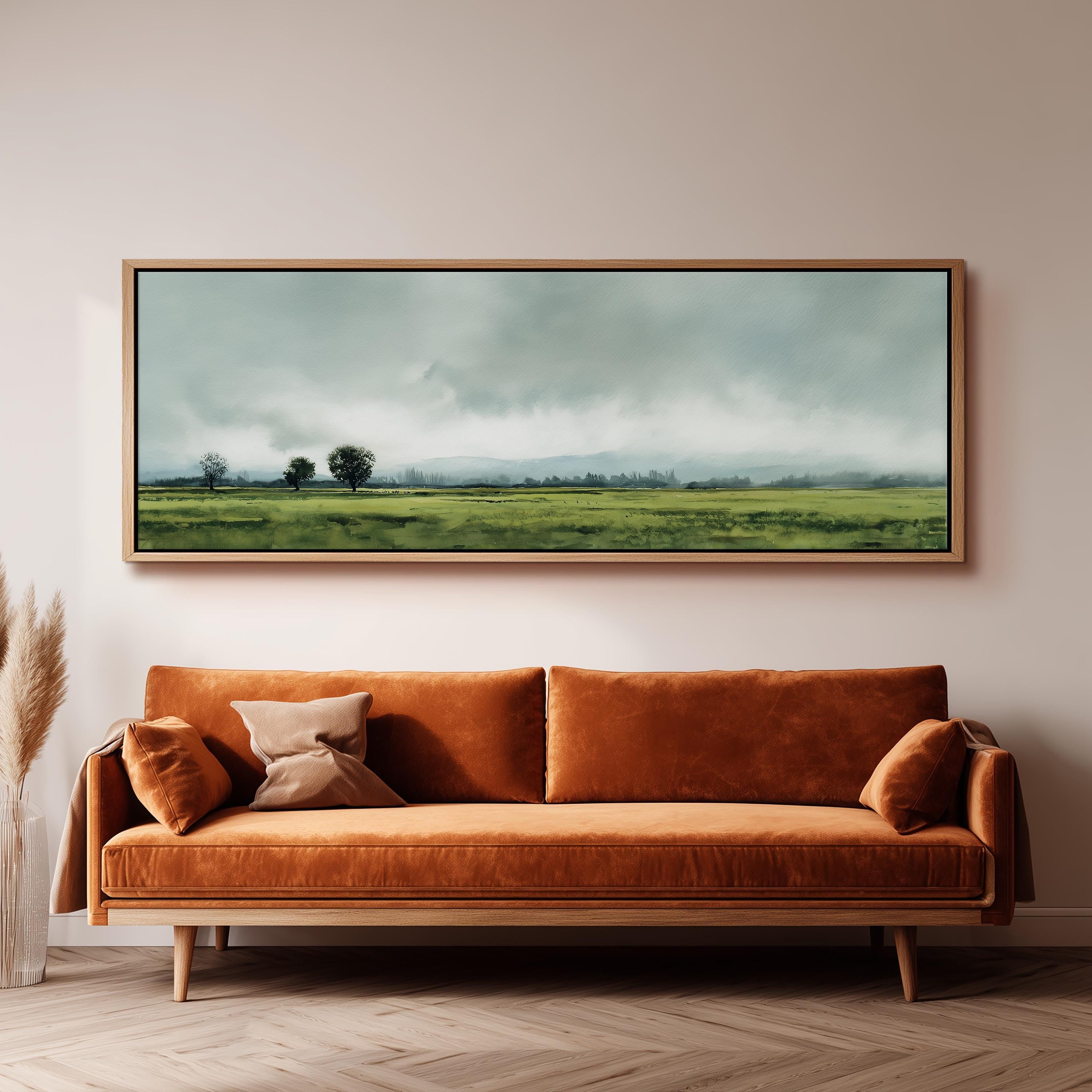a large, framed landscape painting hanging on a wall above a brown sofa with orange cushions.