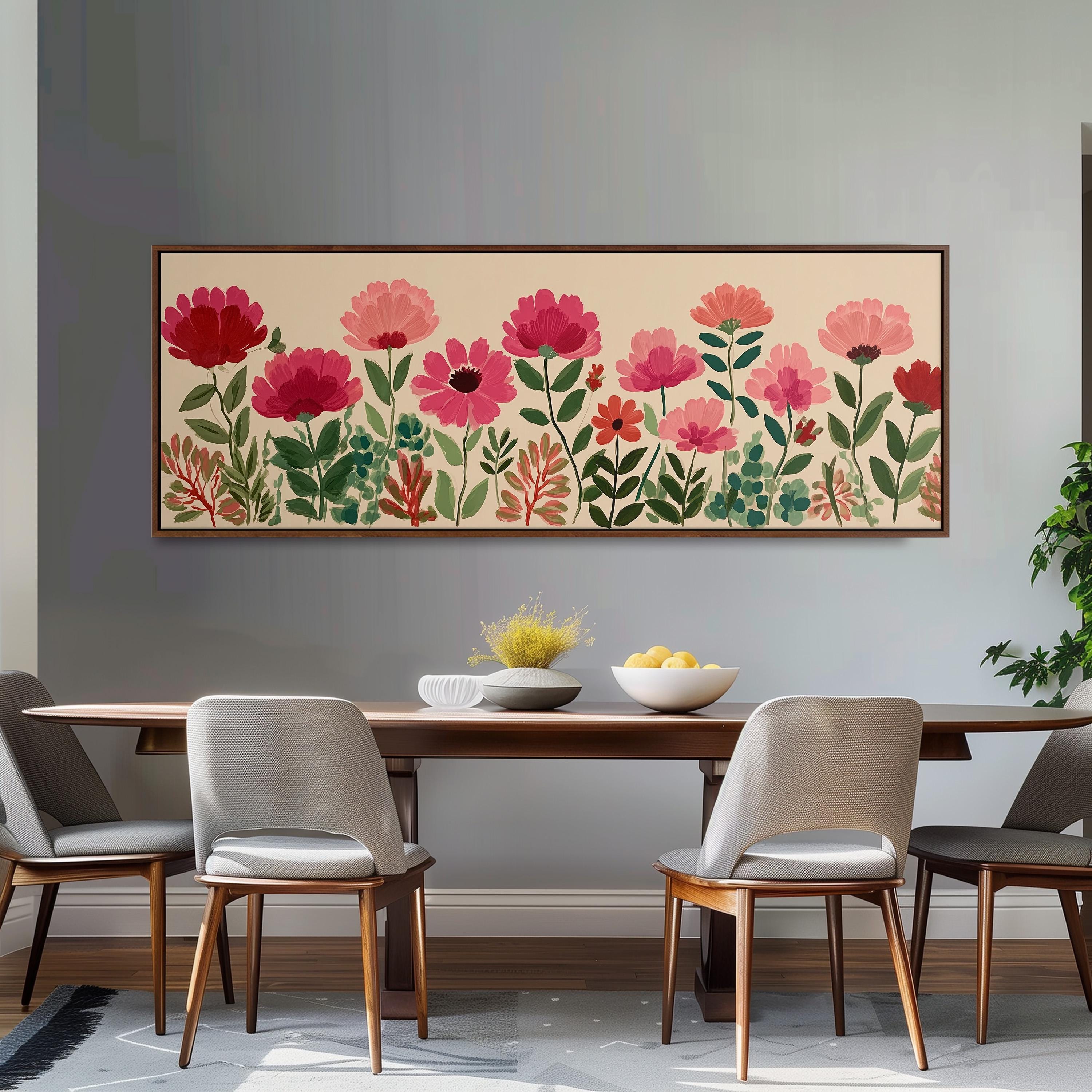 a dining room with a wooden table and four chairs. Above the table, there is a large painting of pink flowers and green leaves. The painting is framed and hangs on the wall. The dining table is set with a bowl of fruit and a vase of flowers.