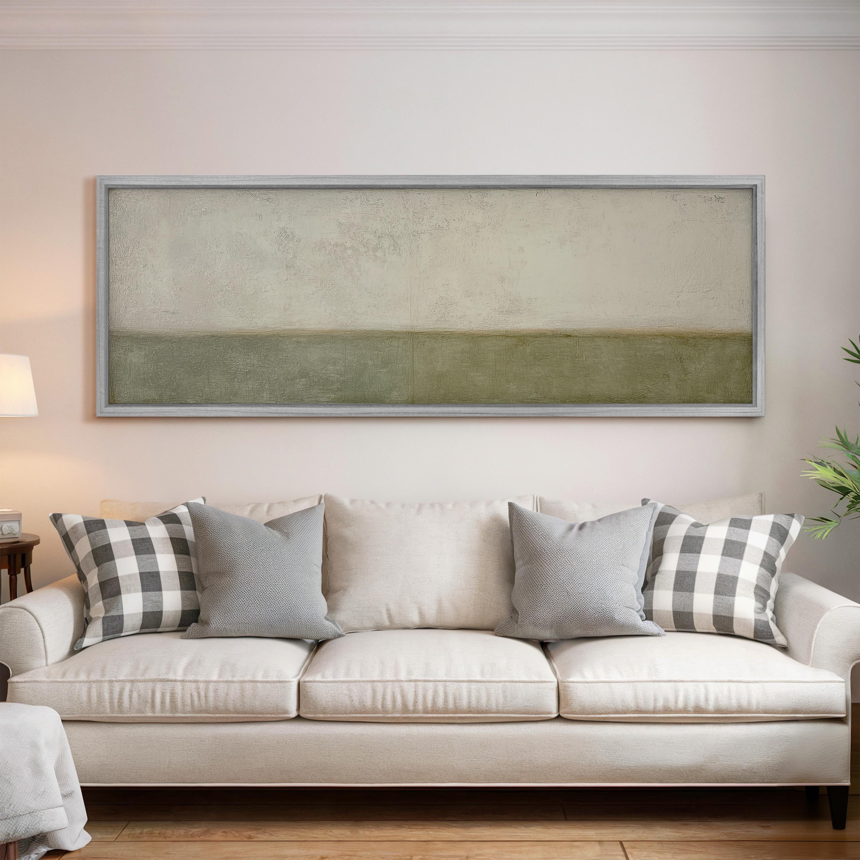 a living room with a white sofa adorned with gray and white patterned throw pillows. Above the sofa, there is a large rectangular painting hanging on the wall.