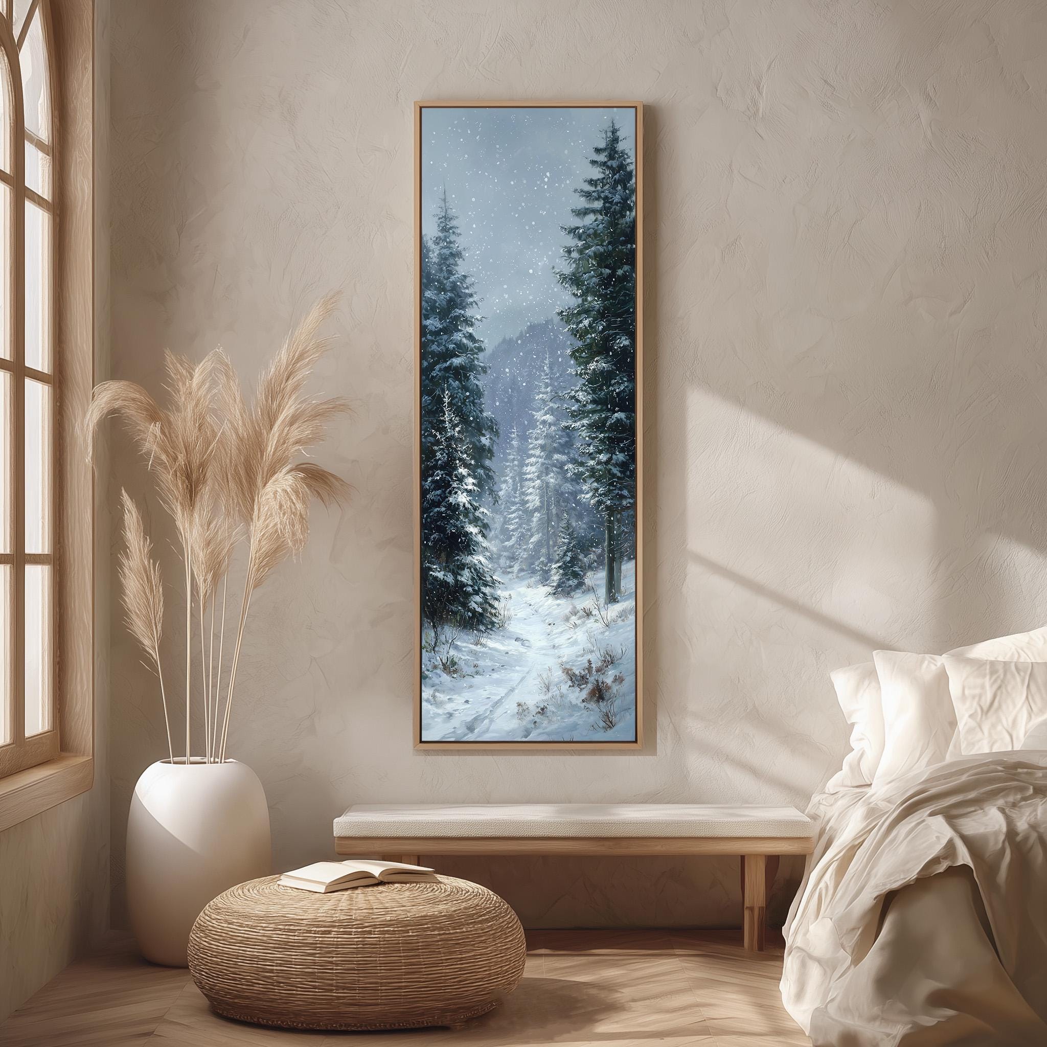 a serene winter scene with a snowy forest, featuring tall trees and a winding path covered in snow. The painting is hung on a wall above a wooden bench, which is positioned in front of a window. The room has a minimalist and cozy atmosphere, with a white bed, a wicker ottoman, and a potted plant adding to the overall aesthetic.