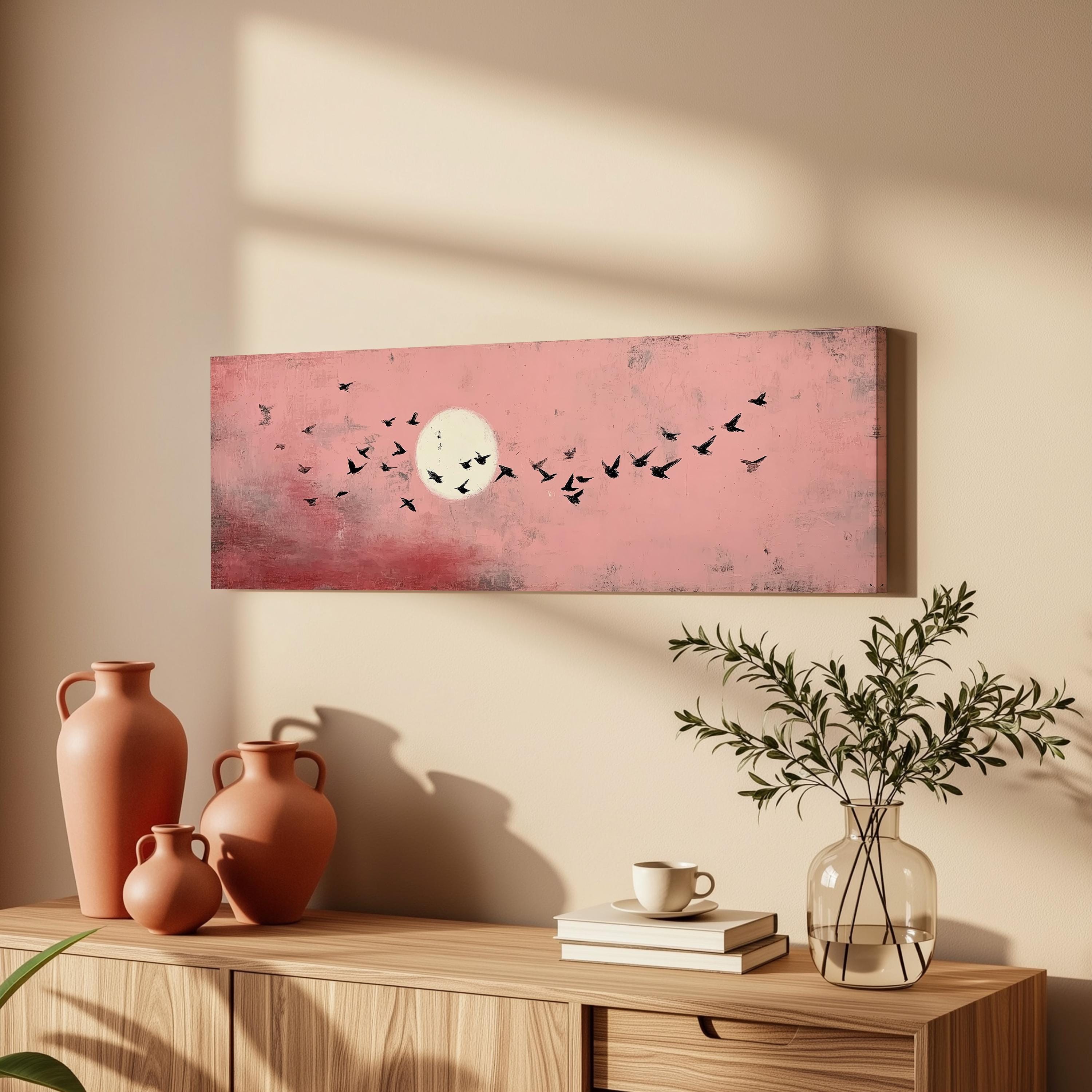 a wooden dresser with a vase of greenery, a stack of books, and a ceramic pitcher. Above the dresser, there is a framed artwork depicting a flock of birds flying against a pink and white background.