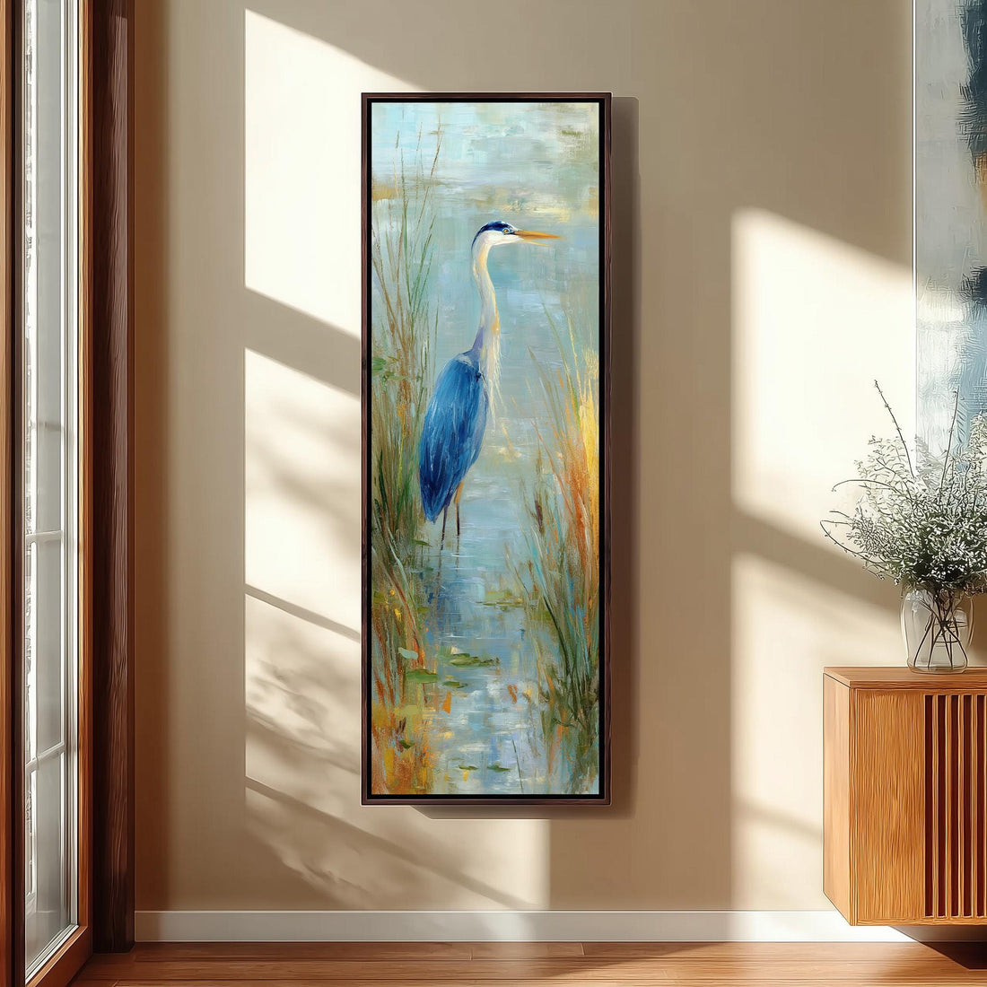 a large painting of a blue heron standing in a pond, surrounded by tall grass and plants. The painting is hung on a wall in a room with a wooden cabinet and a vase of flowers nearby.