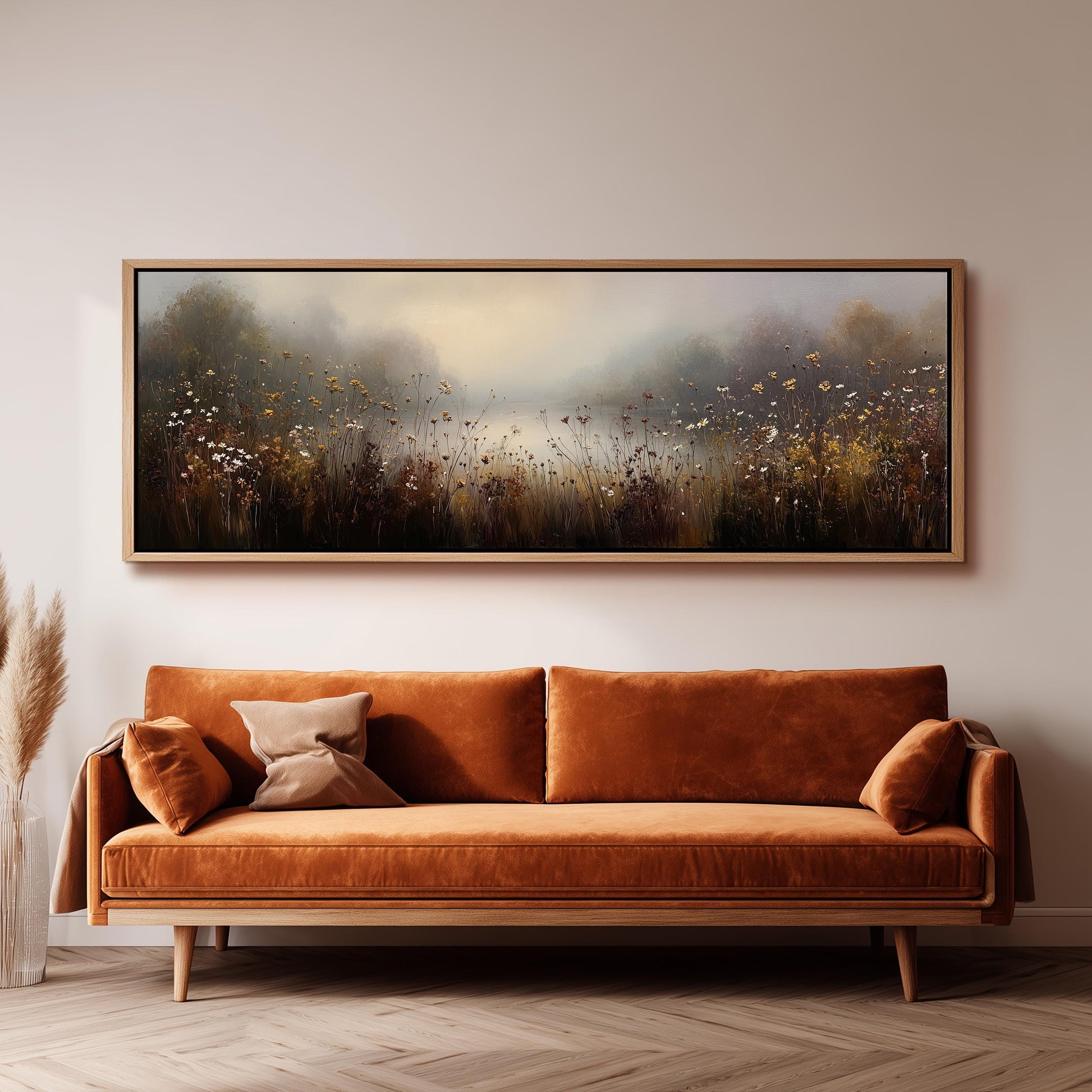 a large, framed painting of a field with flowers hanging on a wall above a brown sofa with orange cushions.