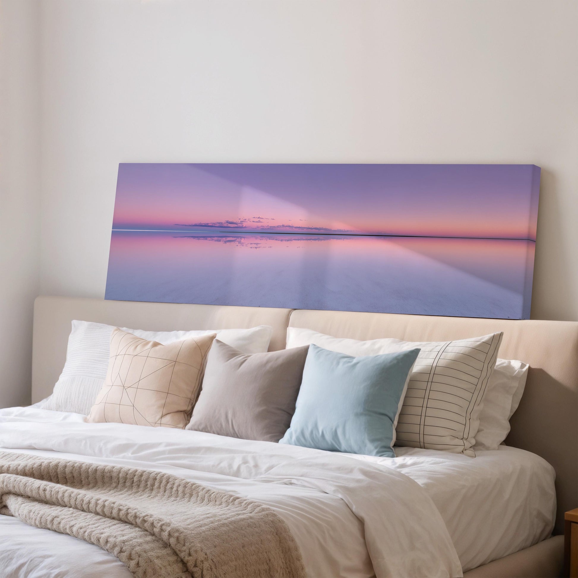a large bedroom with a bed adorned with white linens and a blue pillow. Above the bed, there is a large canvas print depicting a serene sunset over a body of water, creating a peaceful and calming atmosphere in the room.