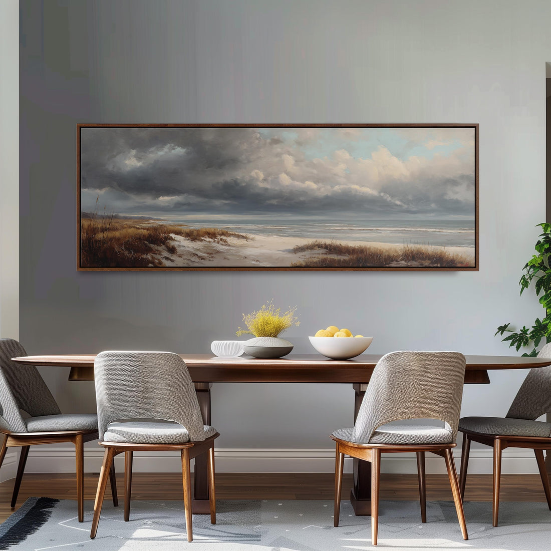 a dining room with a large painting of a beach scene hanging on the wall above a wooden dining table with four chairs. The painting depicts a cloudy sky over the beach, with a body of water visible in the distance. The table is set with a bowl of fruit and a vase of flowers, creating a cozy and inviting atmosphere.
