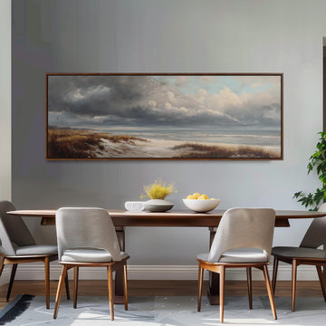 a dining room with a large painting of a beach scene hanging on the wall above a wooden dining table with four chairs. The painting depicts a cloudy sky over the beach, with a body of water visible in the distance. The table is set with a bowl of fruit and a vase of flowers, creating a cozy and inviting atmosphere.