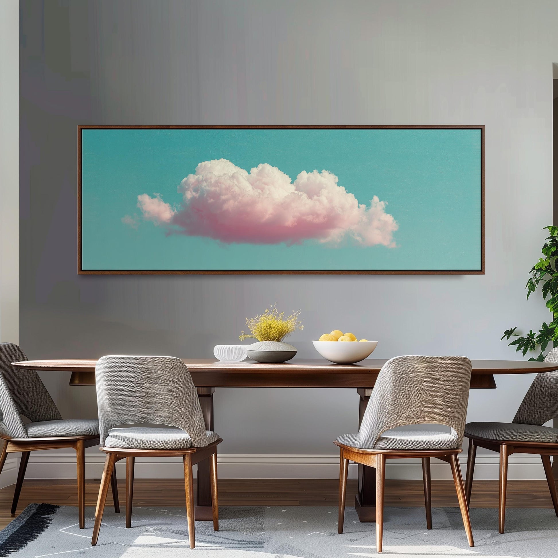 a dining room with a wooden table and four chairs. Above the table, there is a large framed picture of a cloud with pink and white colors. The table is set with a bowl of fruit and a vase of flowers.